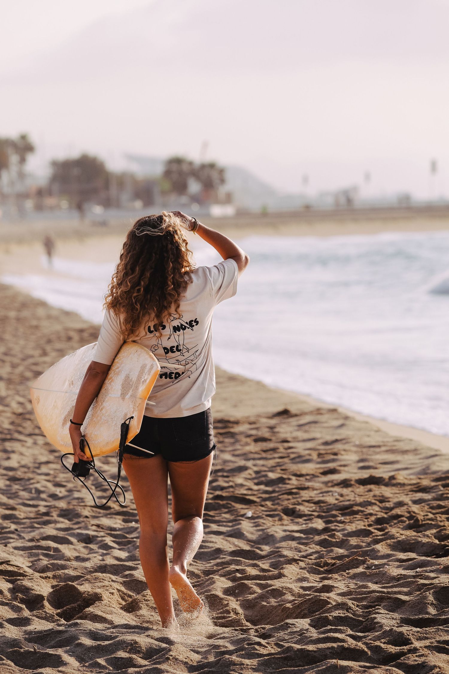 Person carrying surfboard walks along beach at sunset with mountains in background.