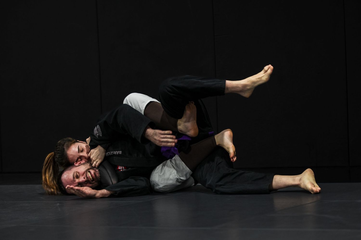 Two martial artists grappling on a dark mat during a Brazilian Jiu-Jitsu training session.