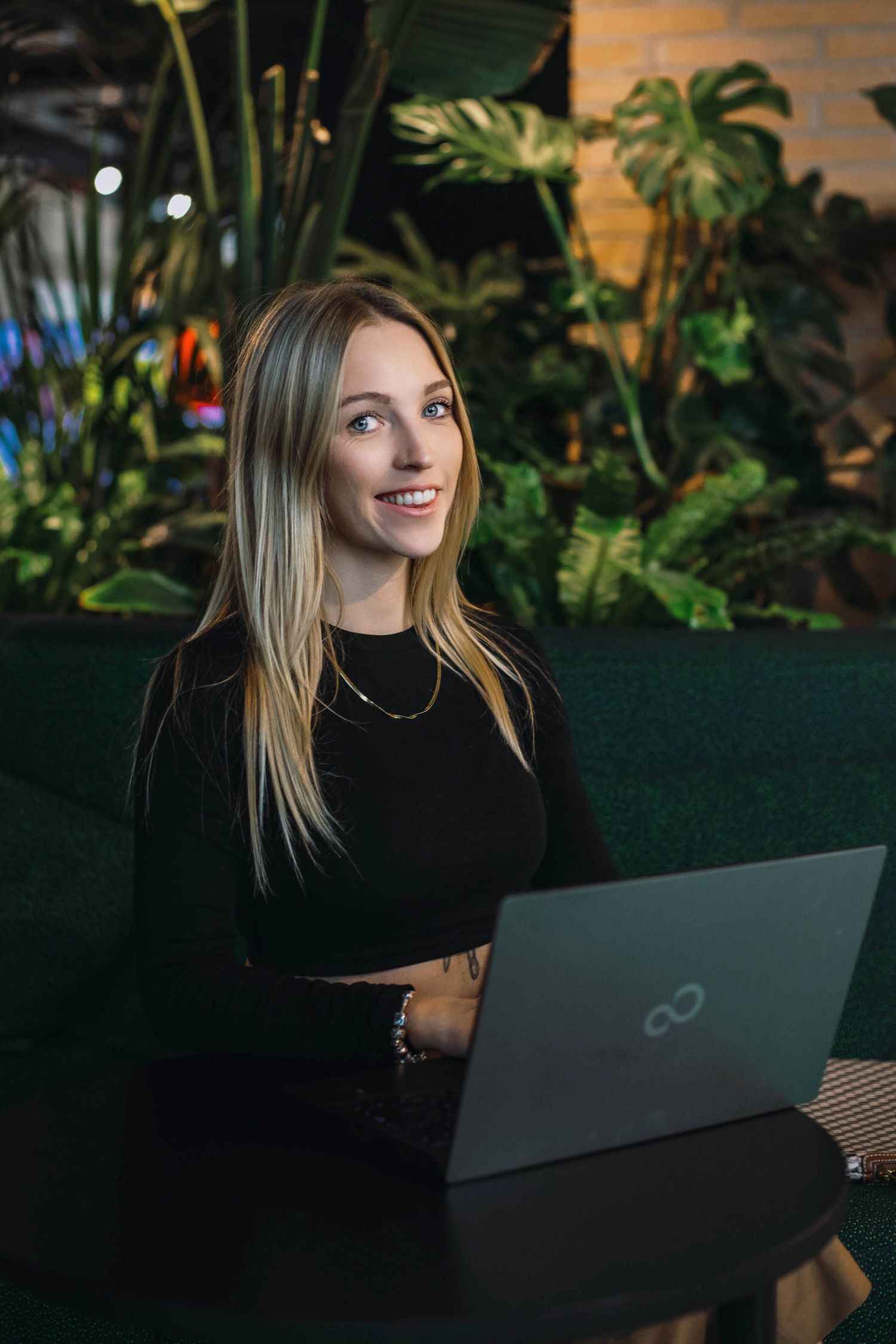 Professional working on laptop in dark setting with tropical plants in background.