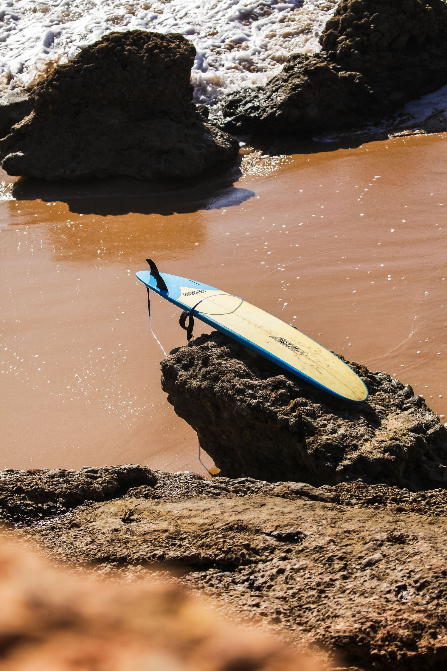 A surfboard rests against rocky outcrops on a beach with reddish-brown sand and ocean waves.