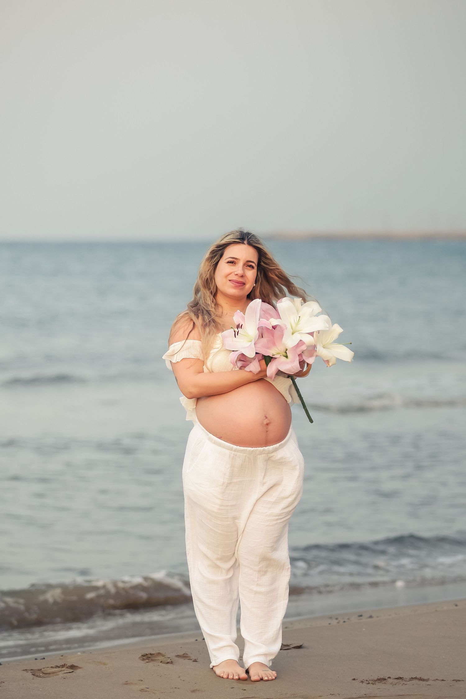 A person in white clothing holds flowers while posing on a beach with ocean in the background.