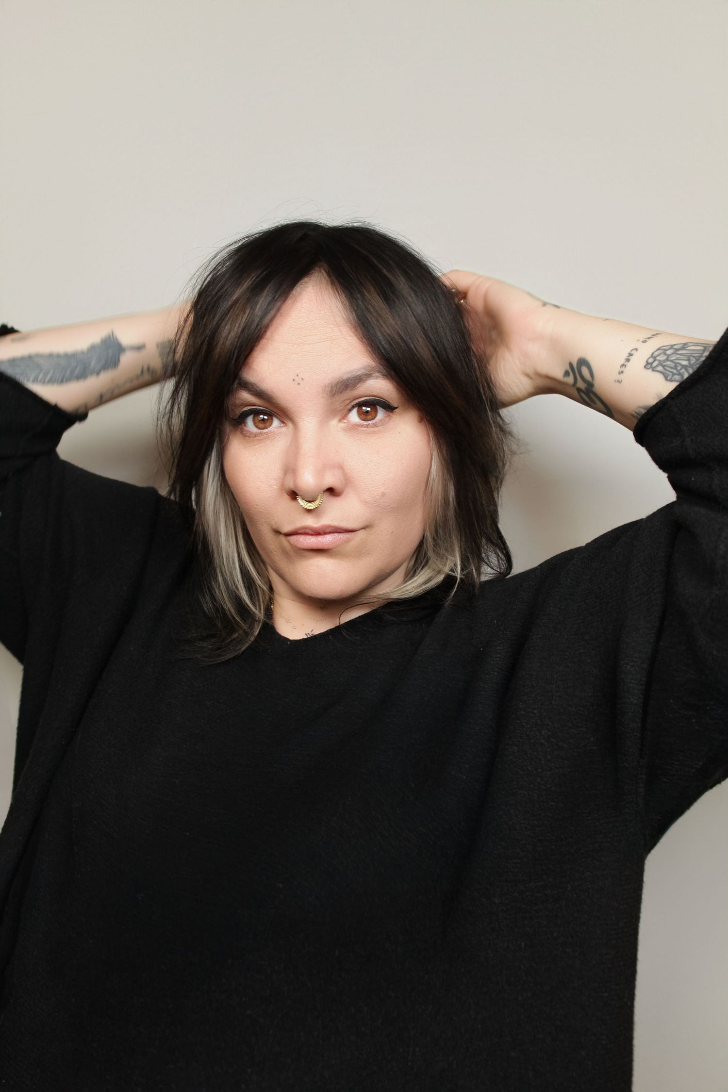 A person wearing a black sweater poses with hands behind head against a light gray background.