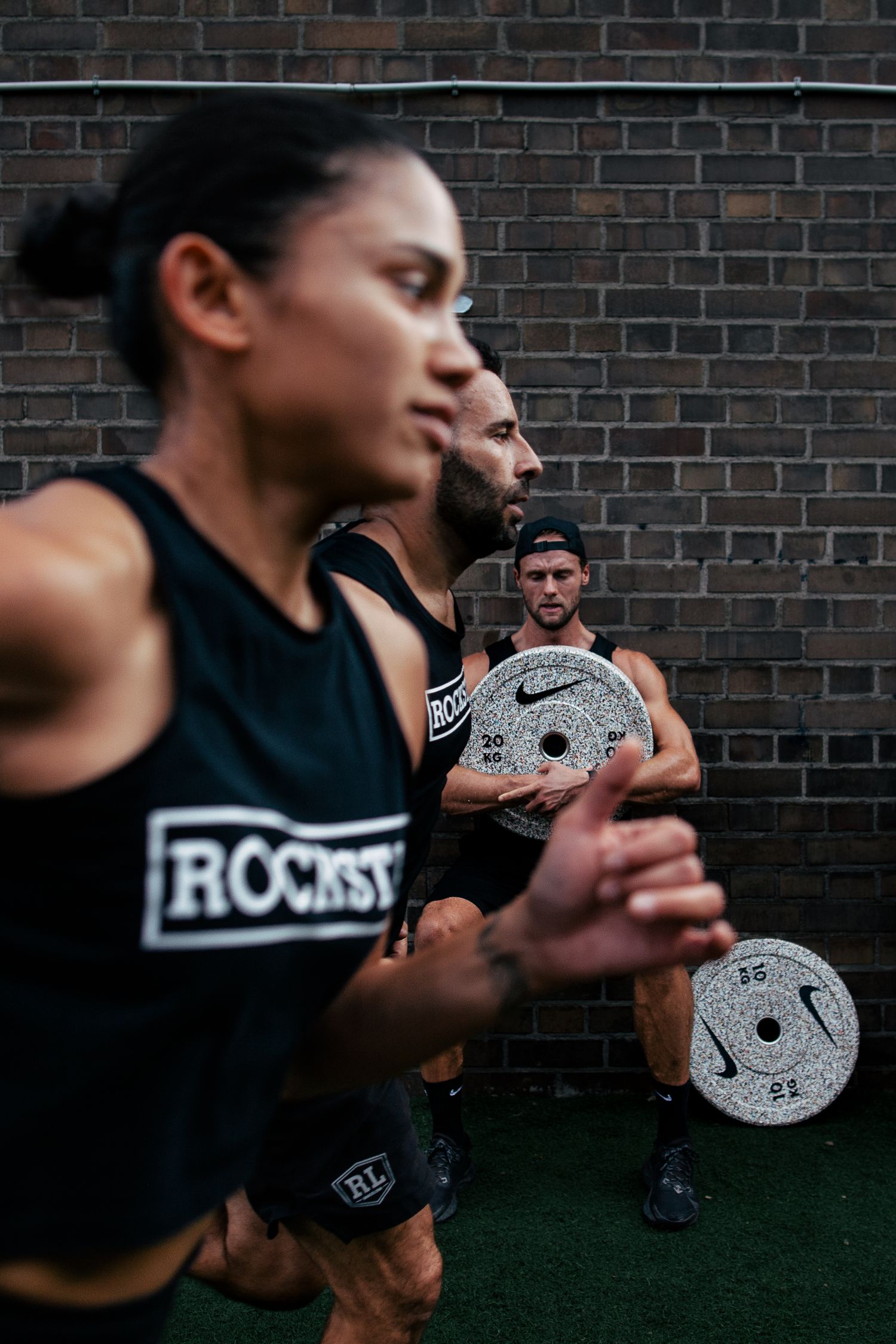 Person in black athletic wear holds speckled weight plates against dark brick wall background.