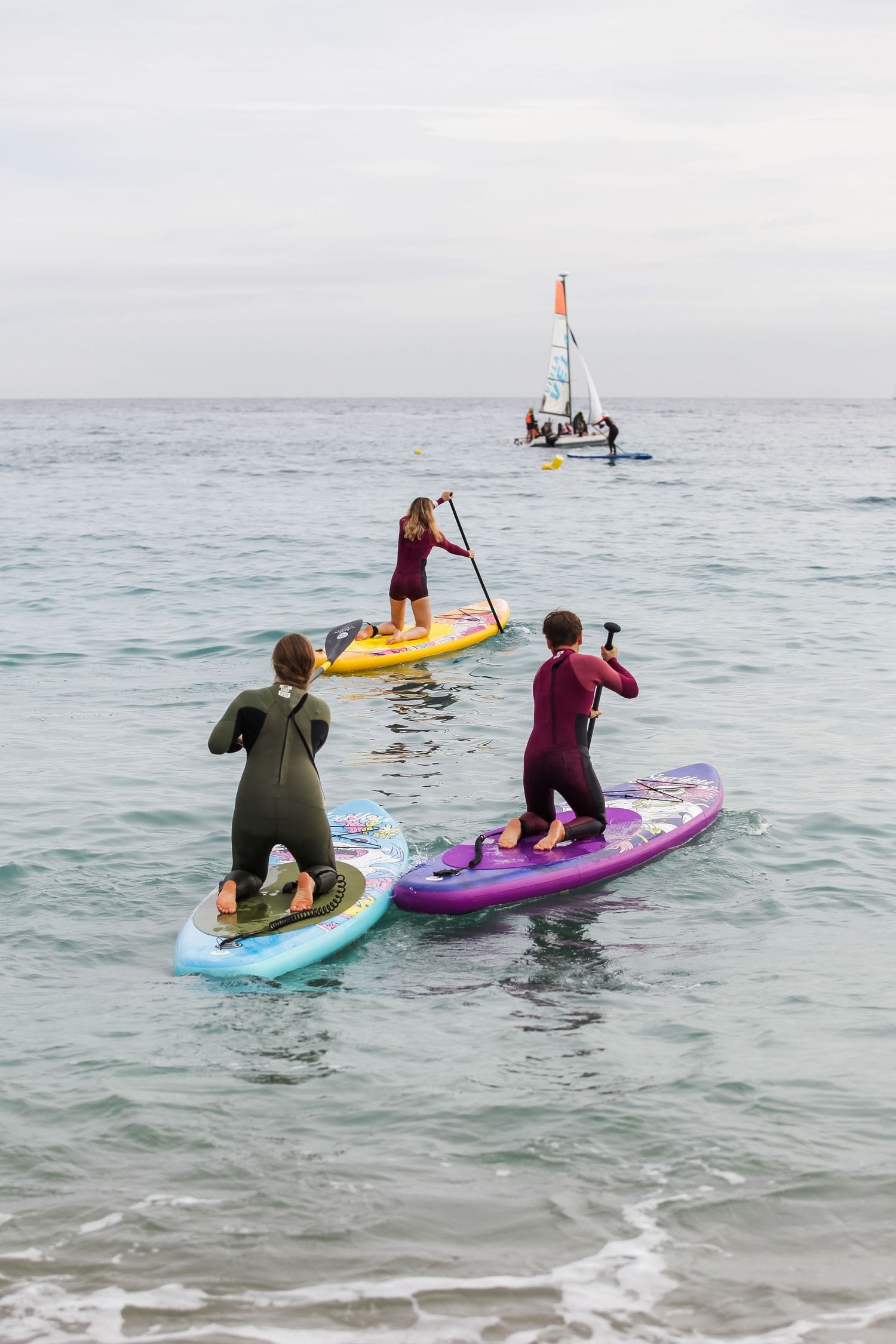 Paddleboarders enjoying water activities with sailboat in background on calm ocean waters.