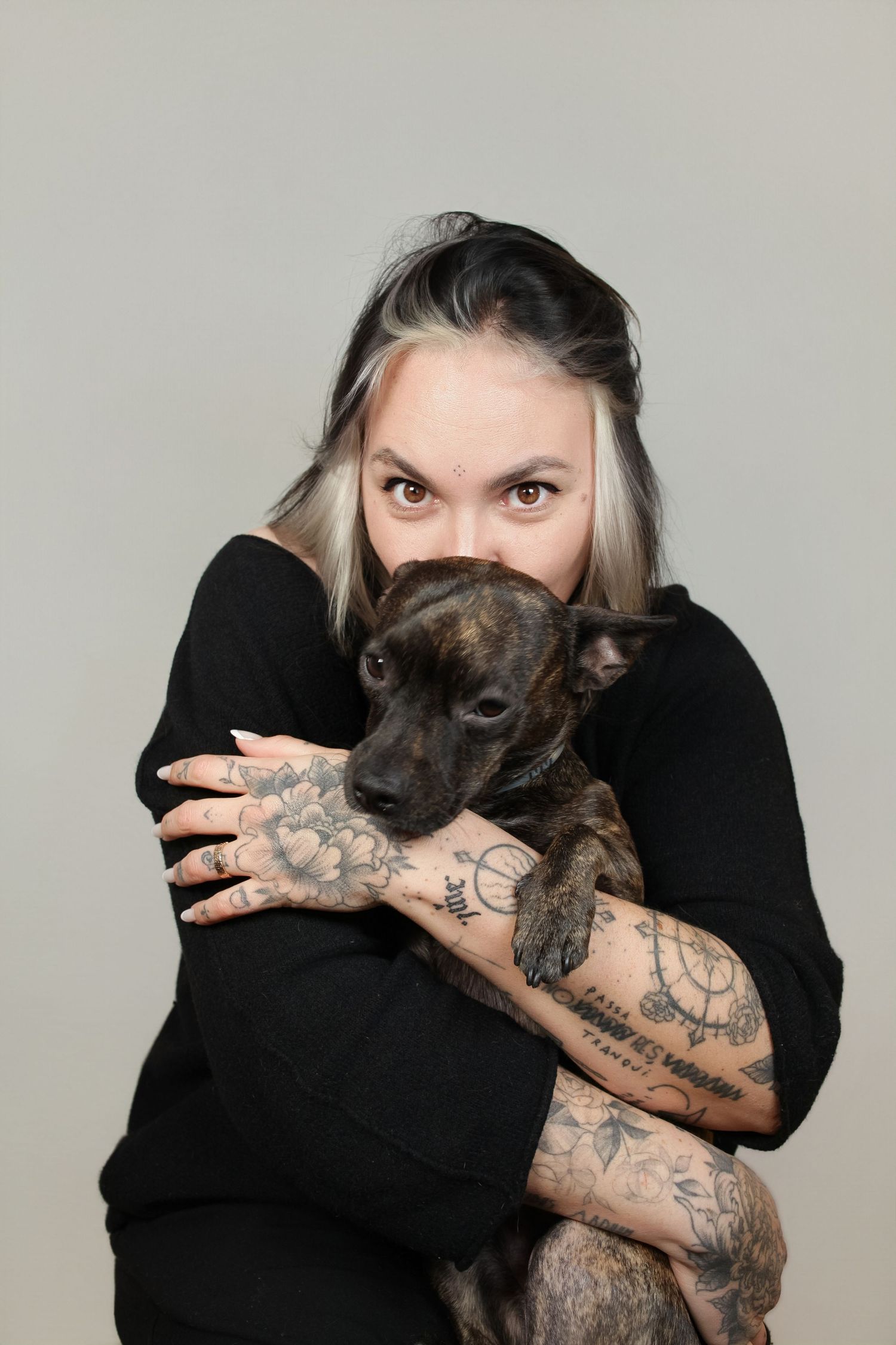 A person wearing black embraces a dark-colored dog with visible tattoos on their arms.