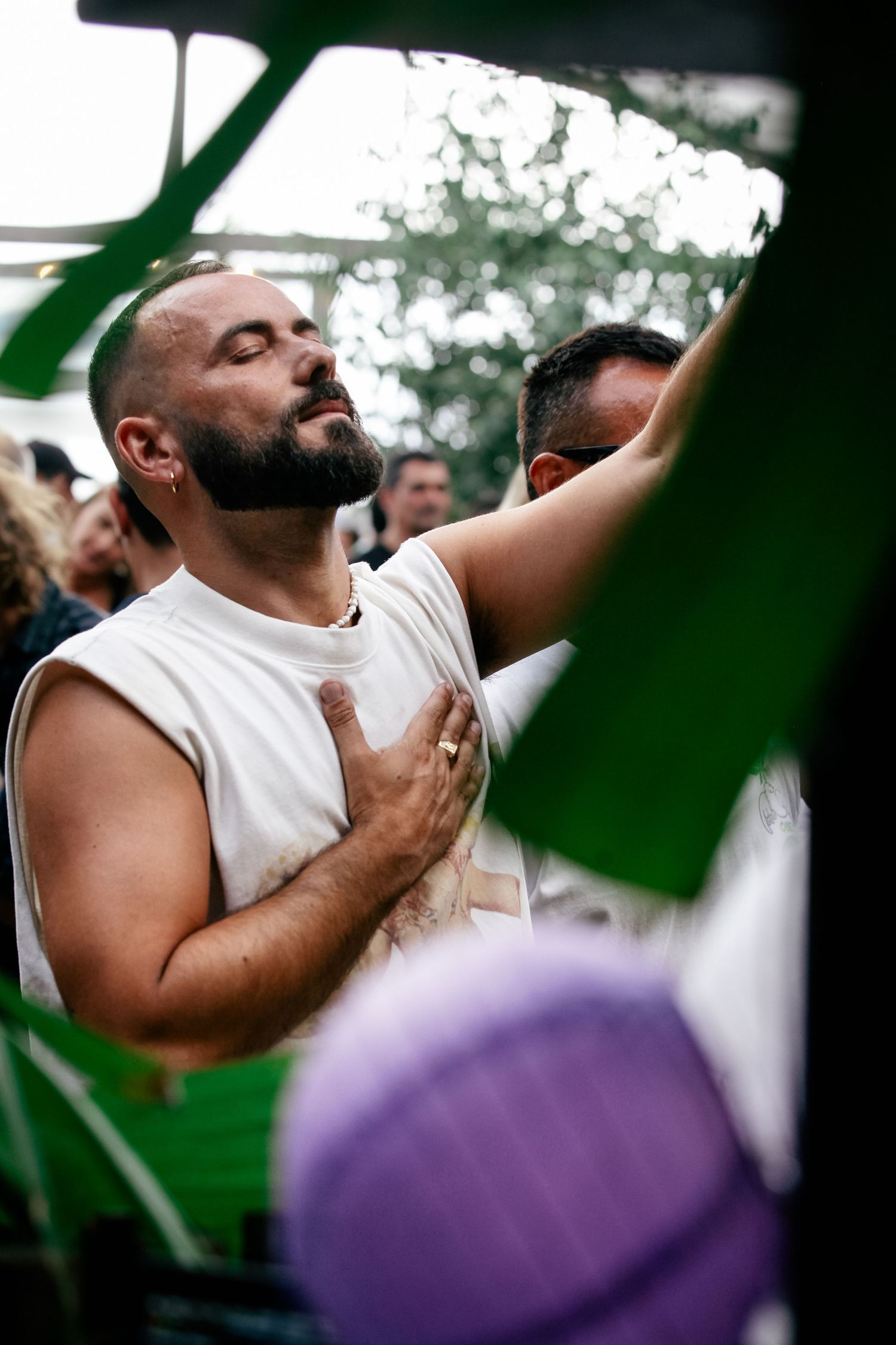 A person in a white sleeveless shirt shown through green foliage at an outdoor gathering.