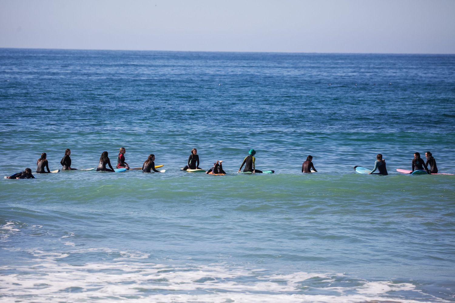 A group of surfers sitting on their boards in a line waiting for waves in blue ocean water.