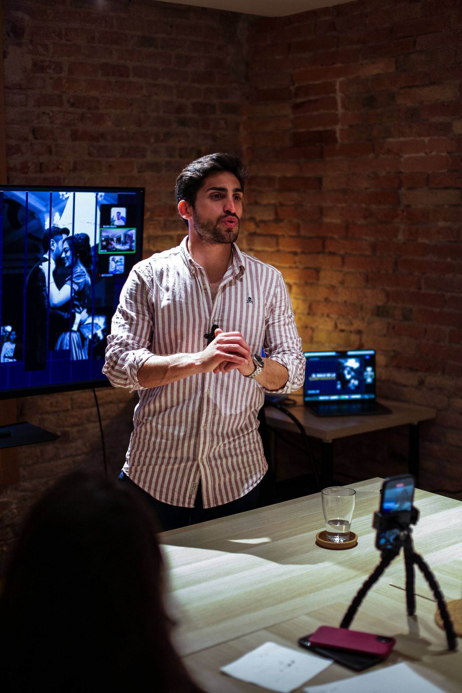 Person gesturing while presenting at a table in a brick-walled room with screens in background.