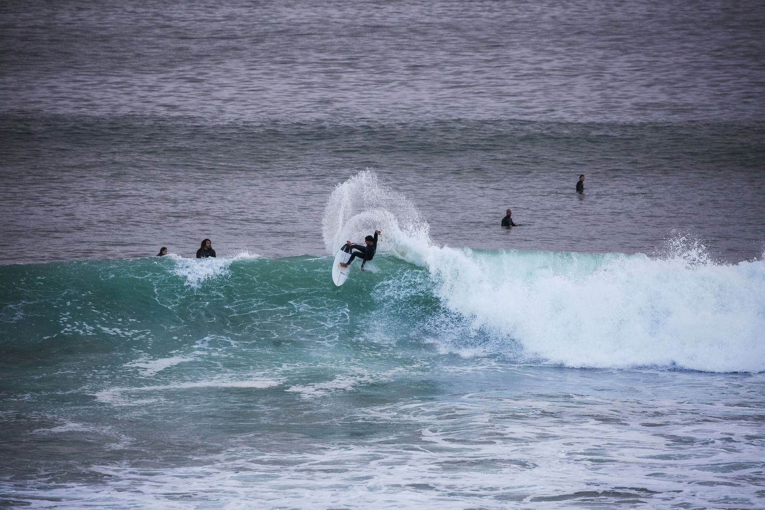 Surfer riding a turquoise wave in stormy ocean conditions with other surfers in the background.