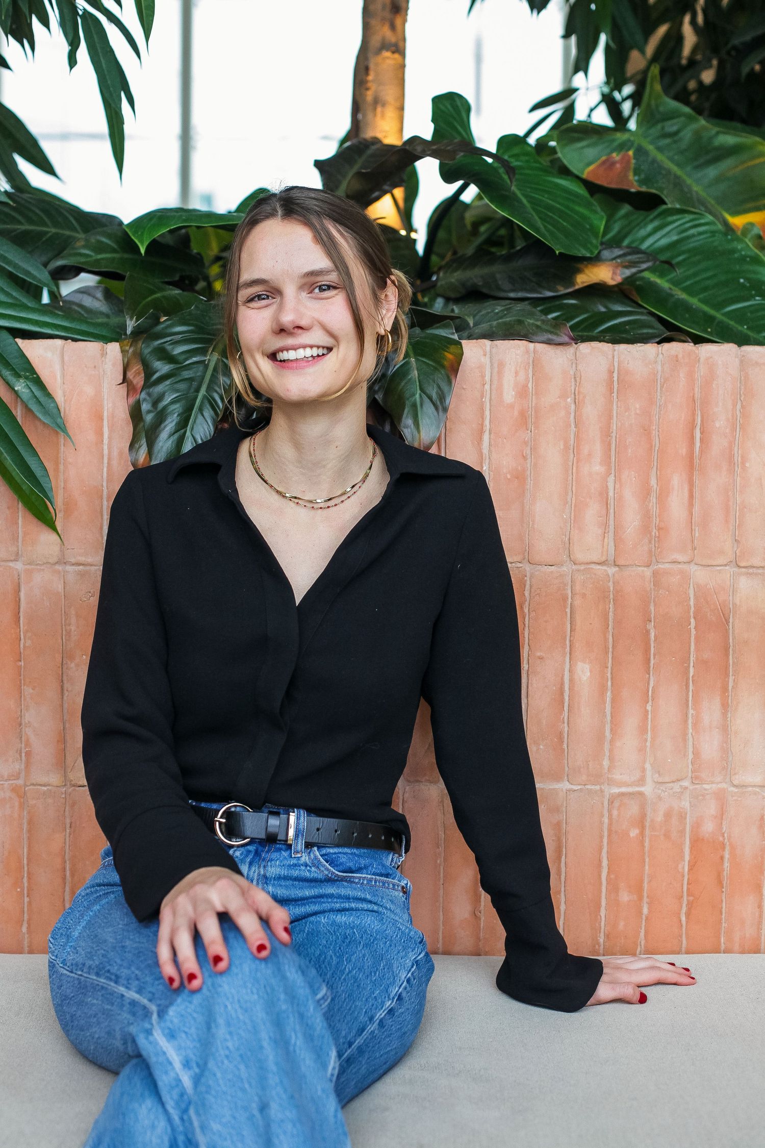 Casual portrait in black top and jeans sitting against brick wall with tropical plants.