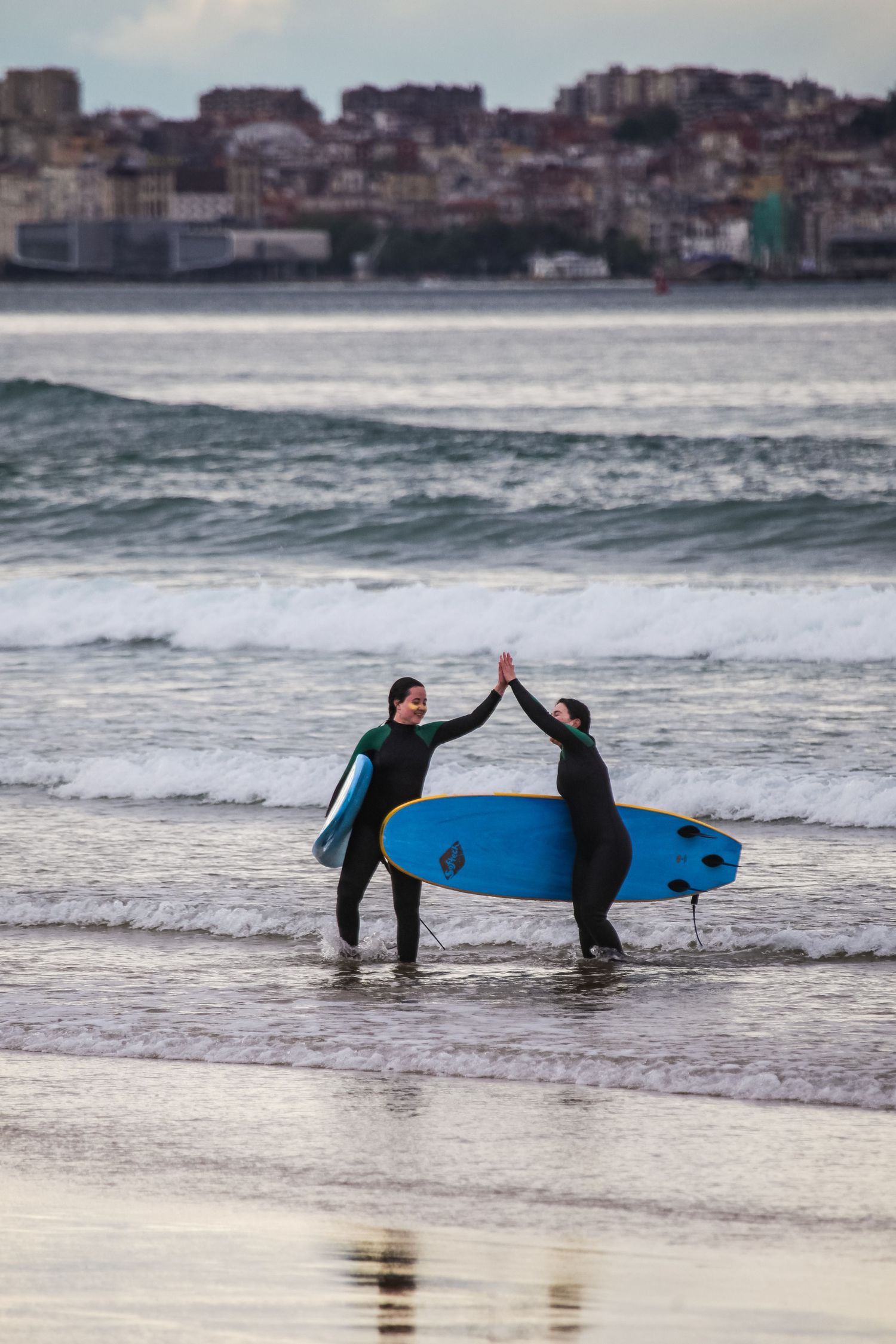Surfers with blue boards high-fiving on a beach with waves and cityscape in background.