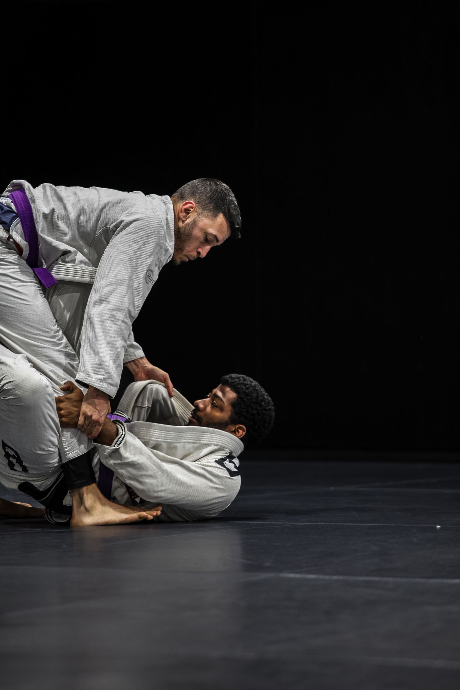Two people practicing Brazilian Jiu-Jitsu grappling moves on a dark mat.