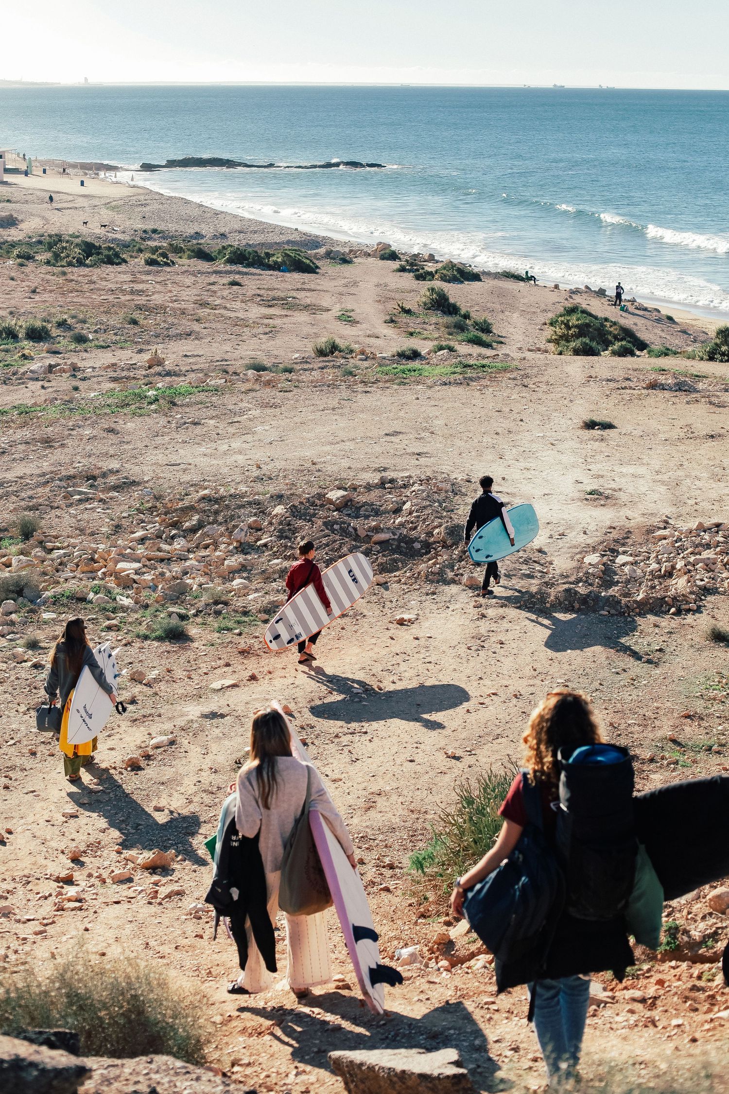 Group walking along a coastal path carrying equipment and bags on a sunny day.