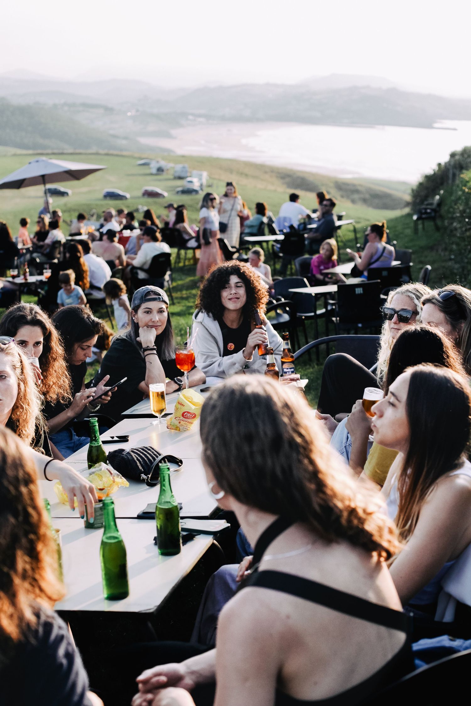 Outdoor gathering with people enjoying food and drinks at picnic tables on a hillside.