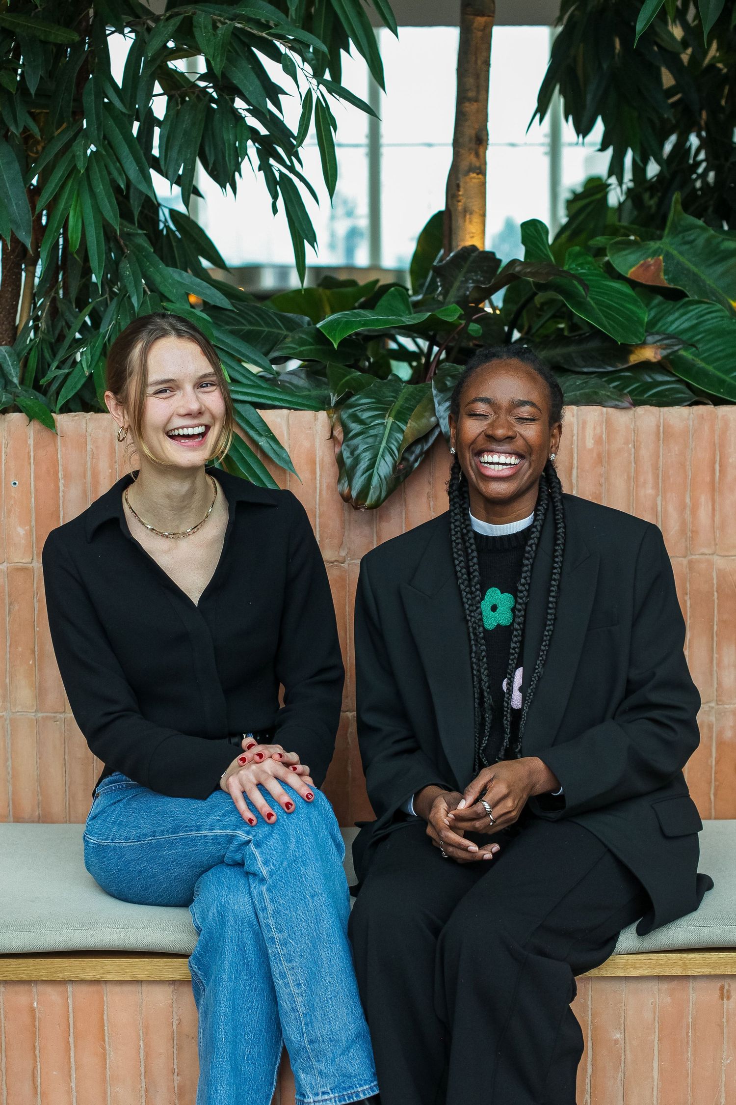 Two people share a joyful moment while sitting on a wooden bench under lush green foliage.