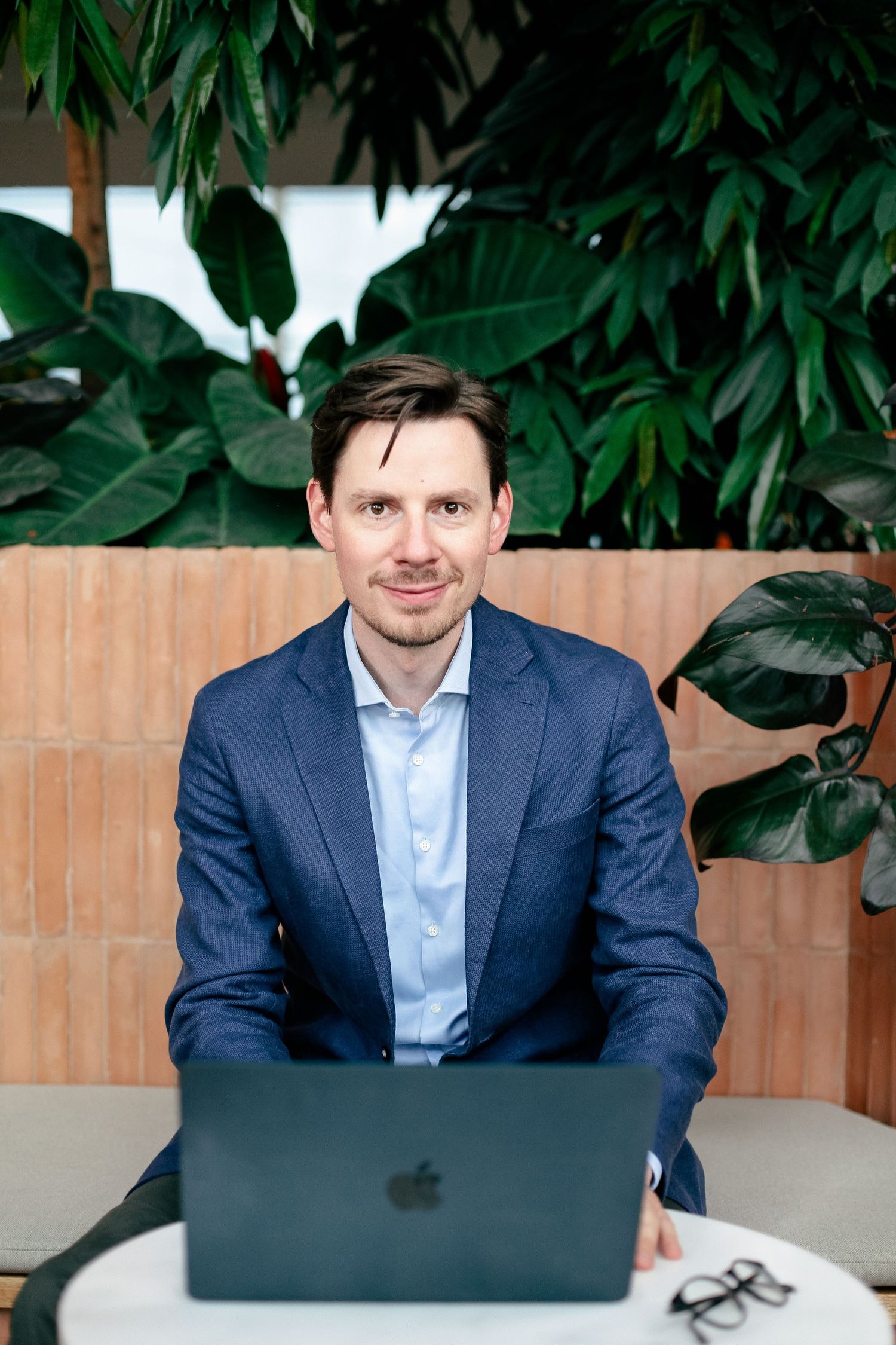 Professional headshot of someone in business attire working on laptop with plants in background.