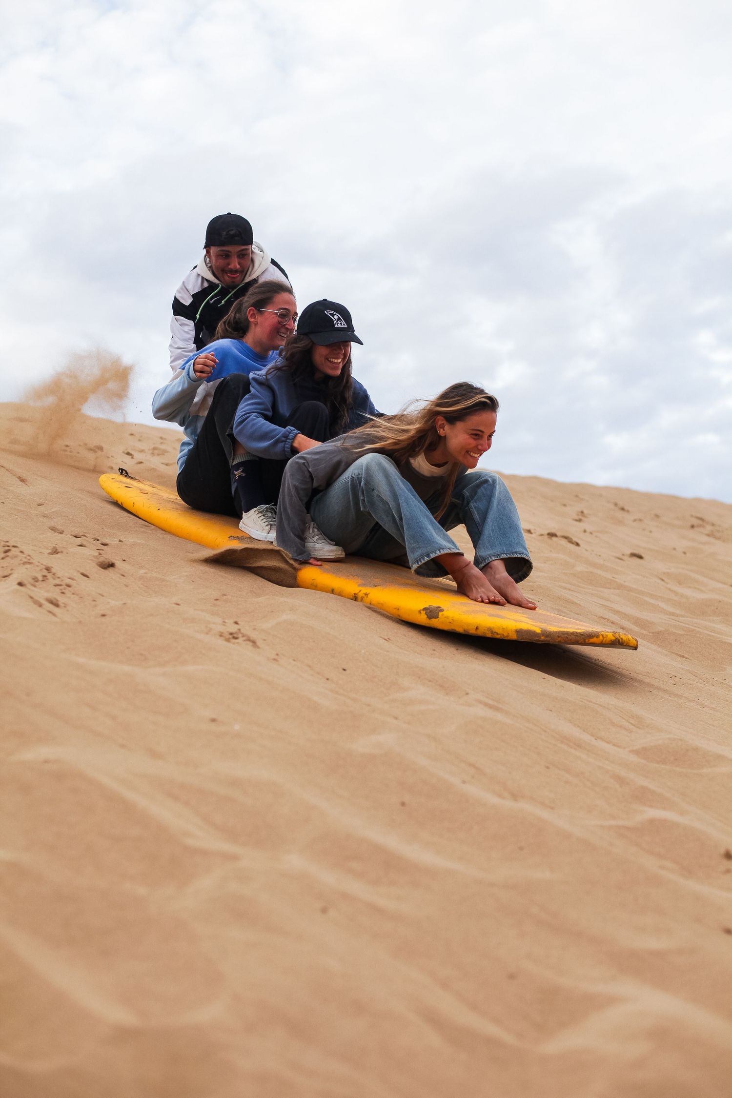 People sandboarding down a steep sand dune on a yellow board.