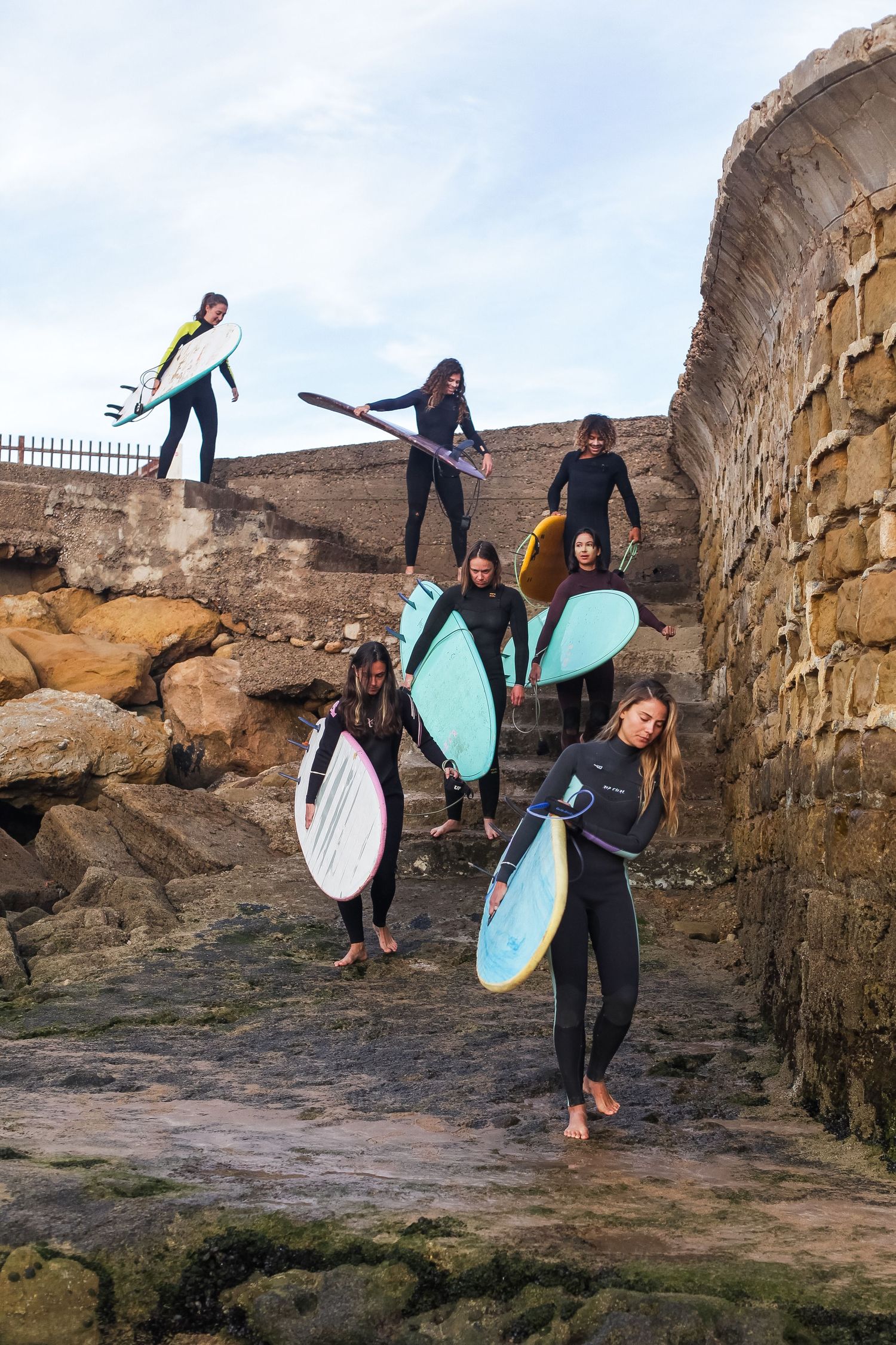 Surfers carrying colorful surfboards down stone steps near an ancient wall.