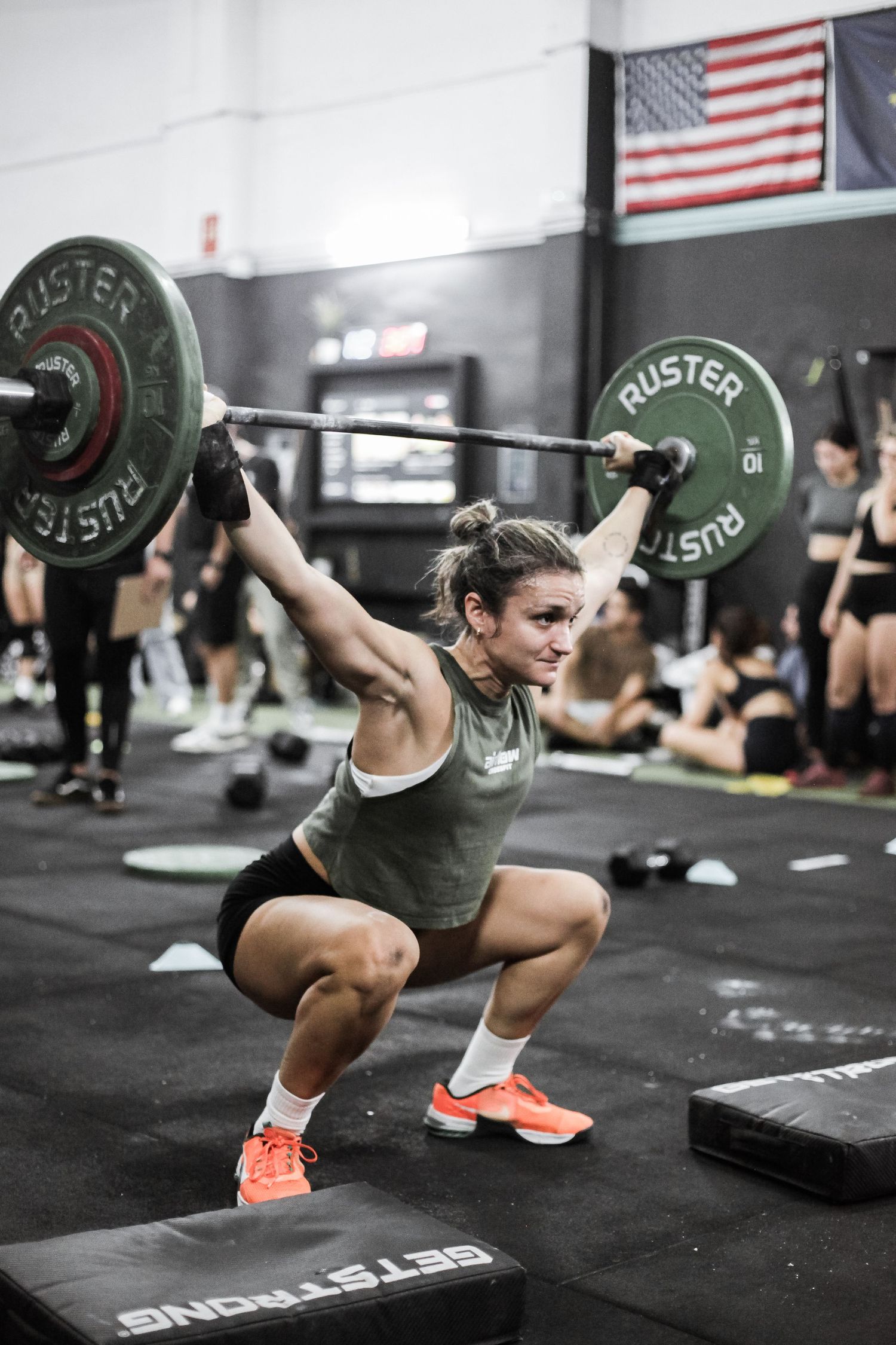 Athlete performing an overhead squat with a barbell in a CrossFit gym with American flag visible.