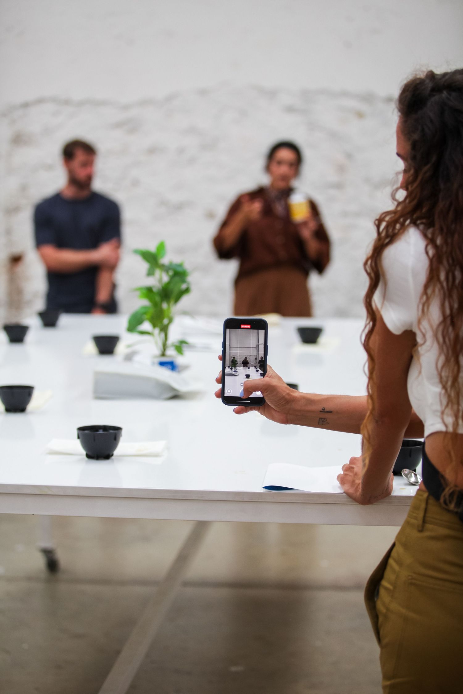 Person taking photo of coffee tasting setup with phone while others stand at white table with plant and black cups.