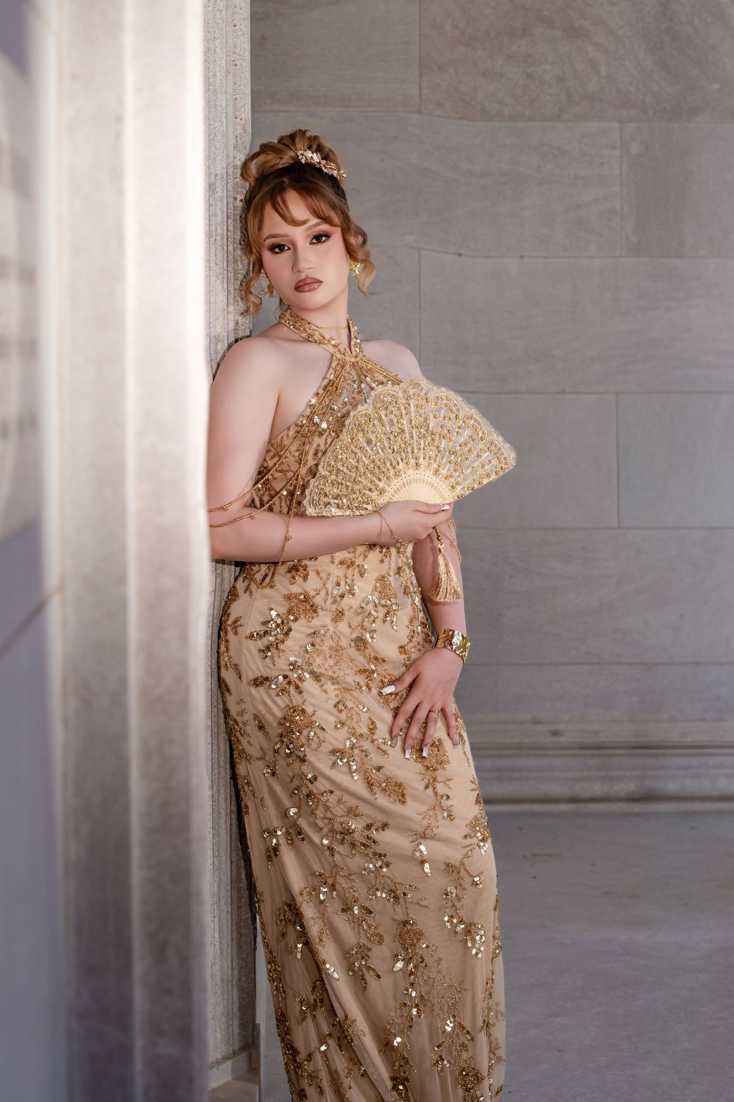 Young woman in gold embroidered halter gown holding ornate fan beside marble column indoors.