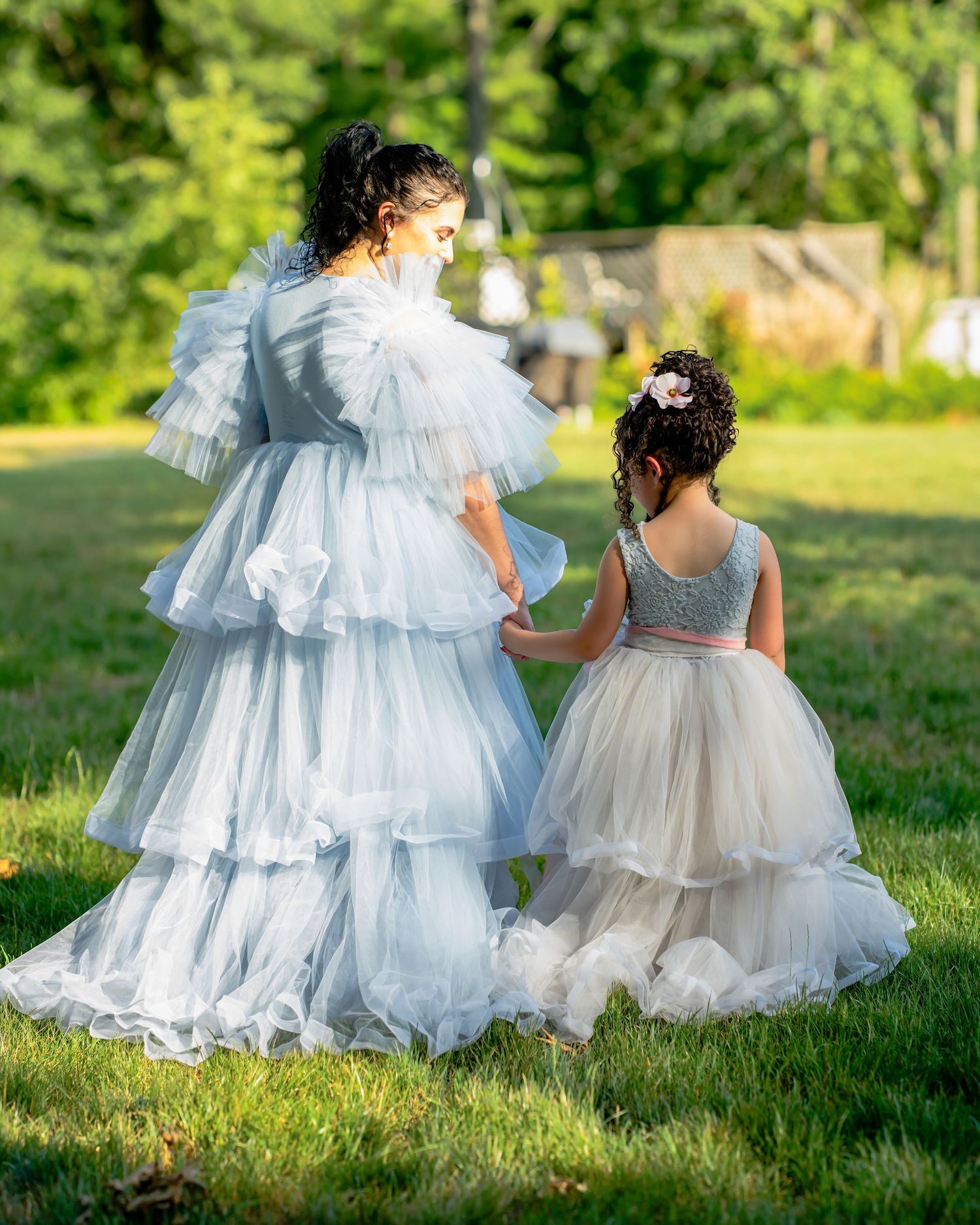 Woman and young girl in layered white tulle gowns walking together on a lush green lawn.