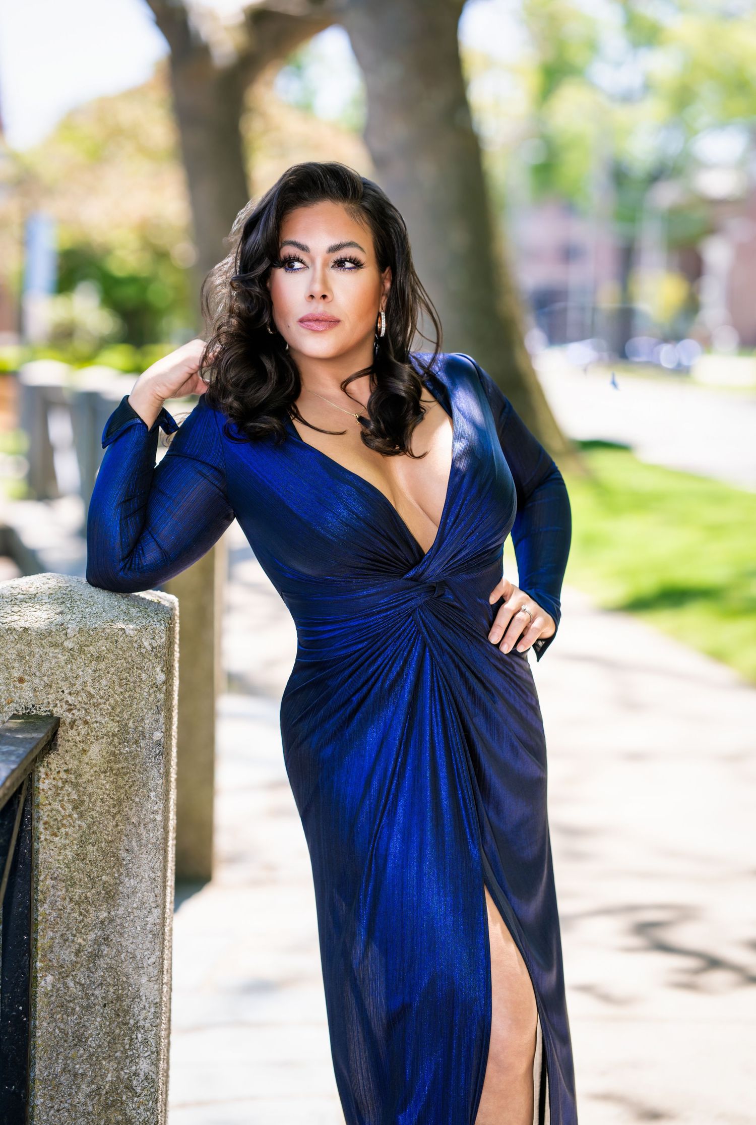 Woman in elegant deep blue long-sleeve gown with plunging neckline posing outdoors on urban sidewalk.