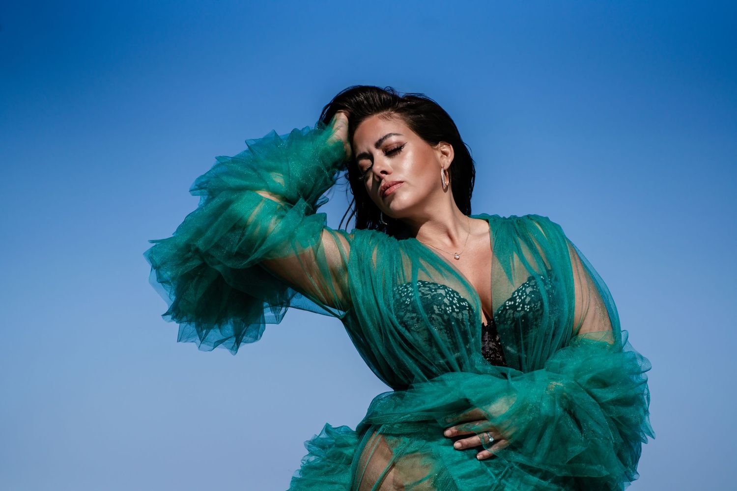 Woman in sheer teal ruffled gown posing dramatically against bright blue sky background.