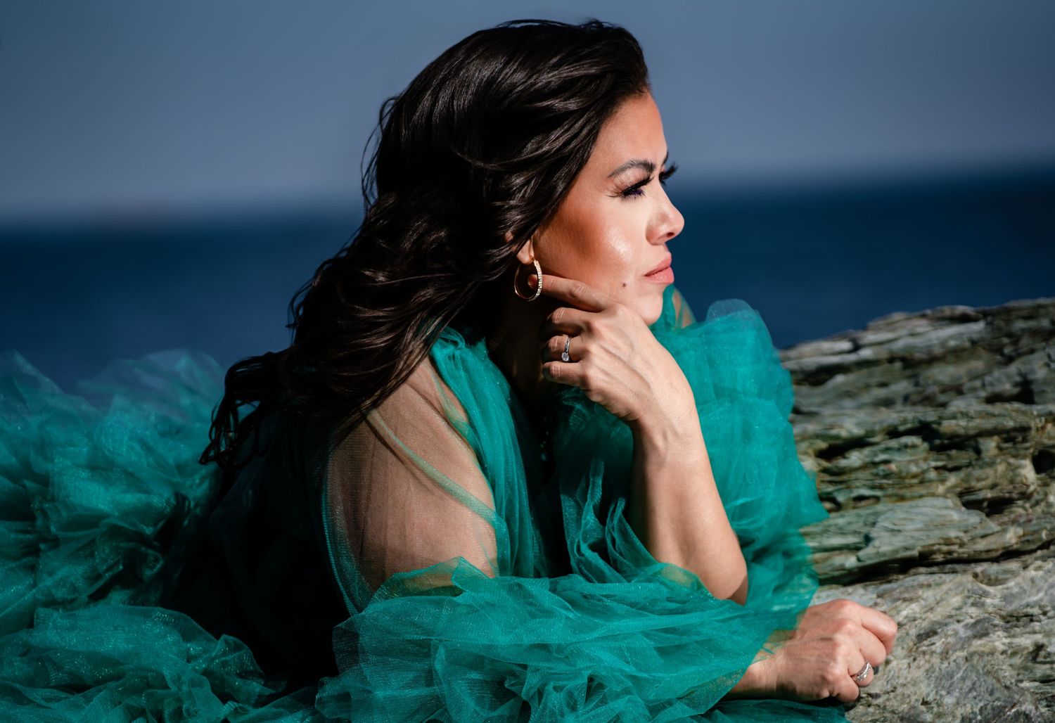 Woman in teal tulle gown with gold earrings resting on rocks near ocean shoreline.