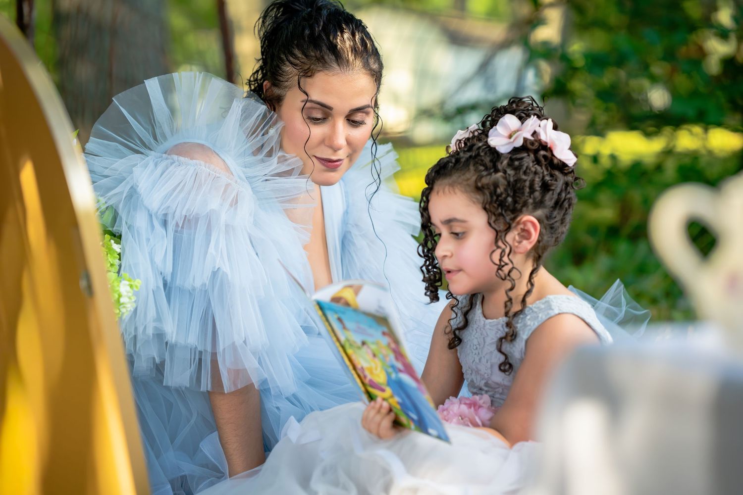 Woman in a blue tulle gown reading a storybook with a young girl in a white princess dress outdoors.