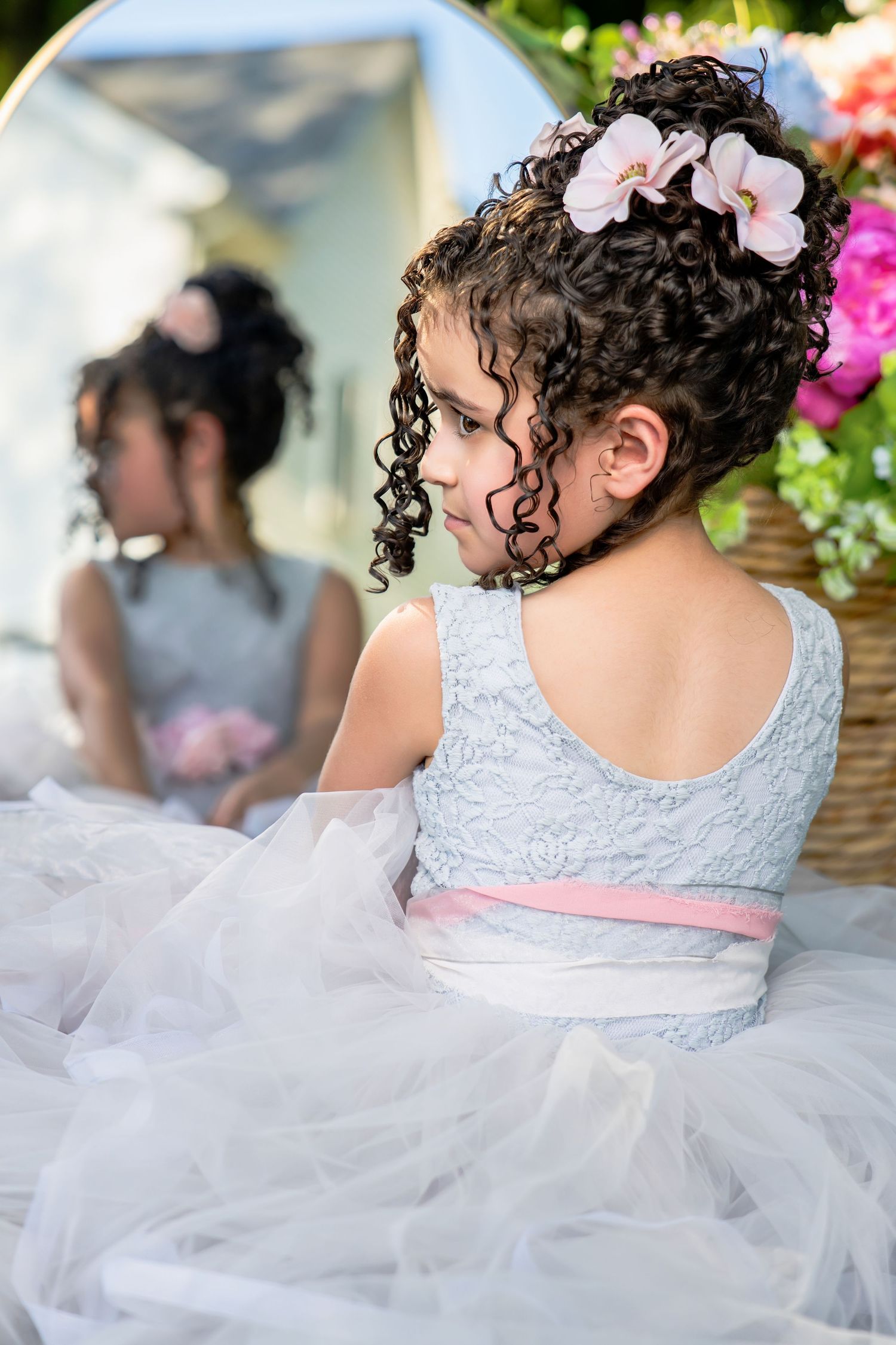 Young girl with curly hair and floral accessories in a white tulle dress sitting near a mirror outdoors.