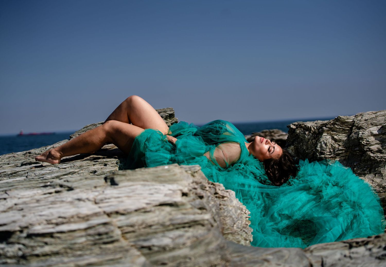 Woman in teal tulle gown poses on rocky coastal cliffs with blue ocean and sky in background.