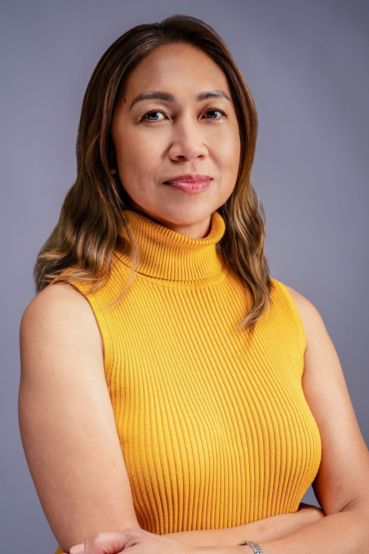Professional portrait of a woman in a yellow turtleneck sweater with arms crossed against a gray background.