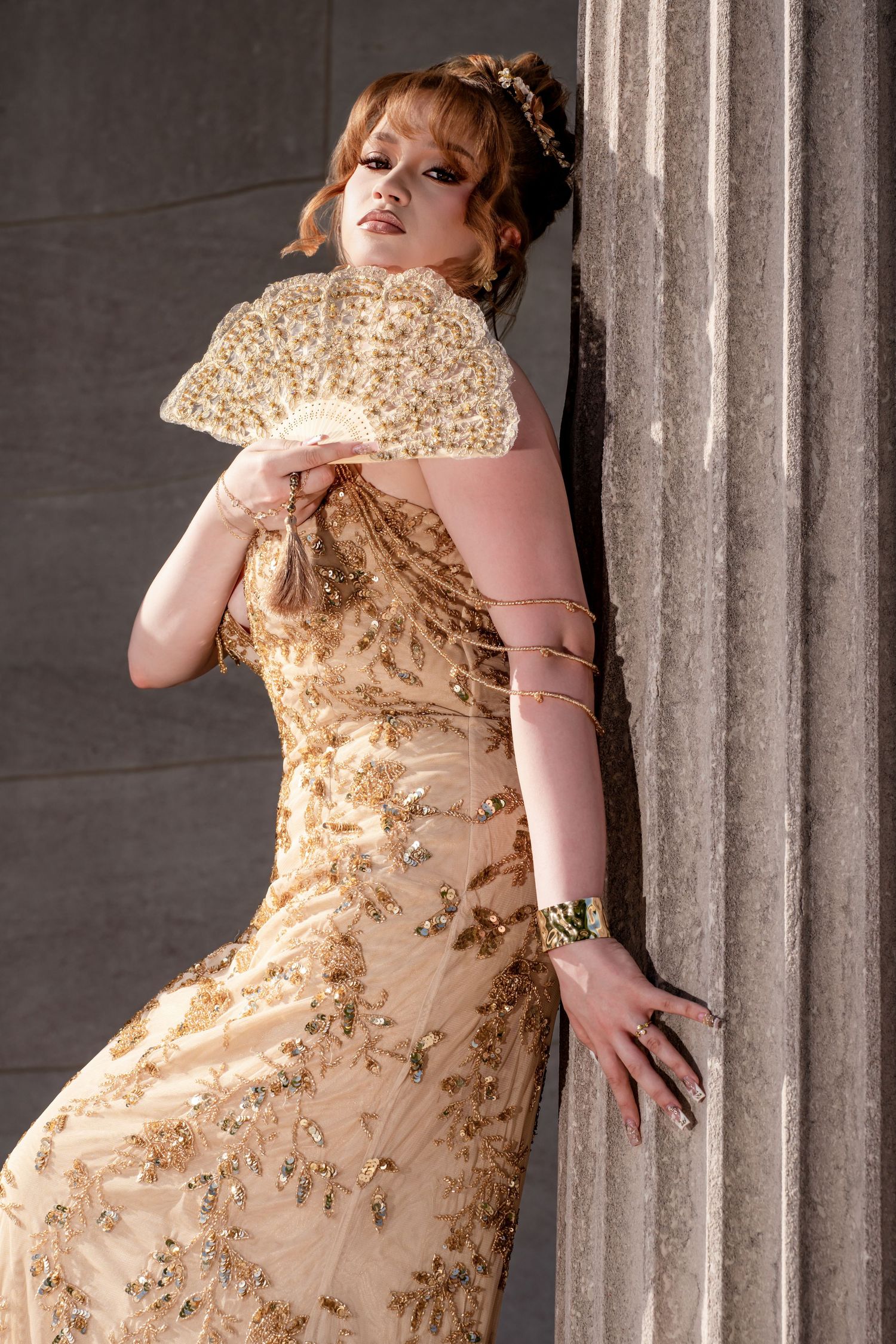 Young woman in gold sequined gown holding decorative fan while leaning against stone pillar.