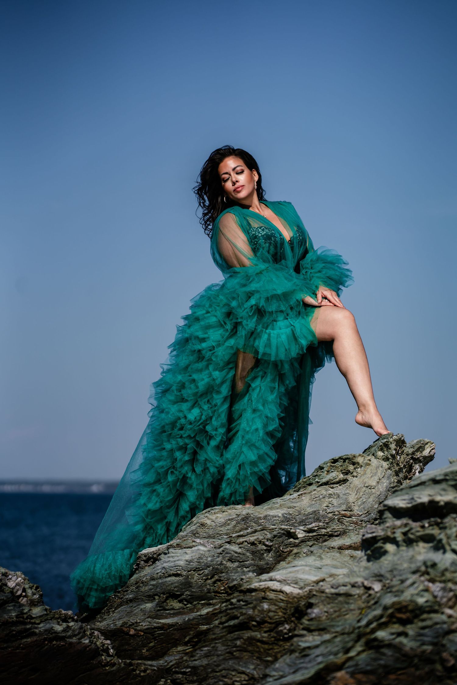 Woman in teal ruffled tulle gown posing barefoot on coastal rocks with ocean backdrop.