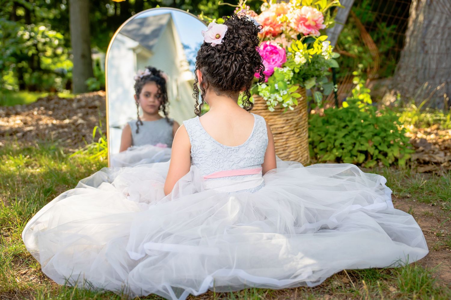 Young girl in a white tulle ball gown sitting in front of a mirror surrounded by flowers outdoors.