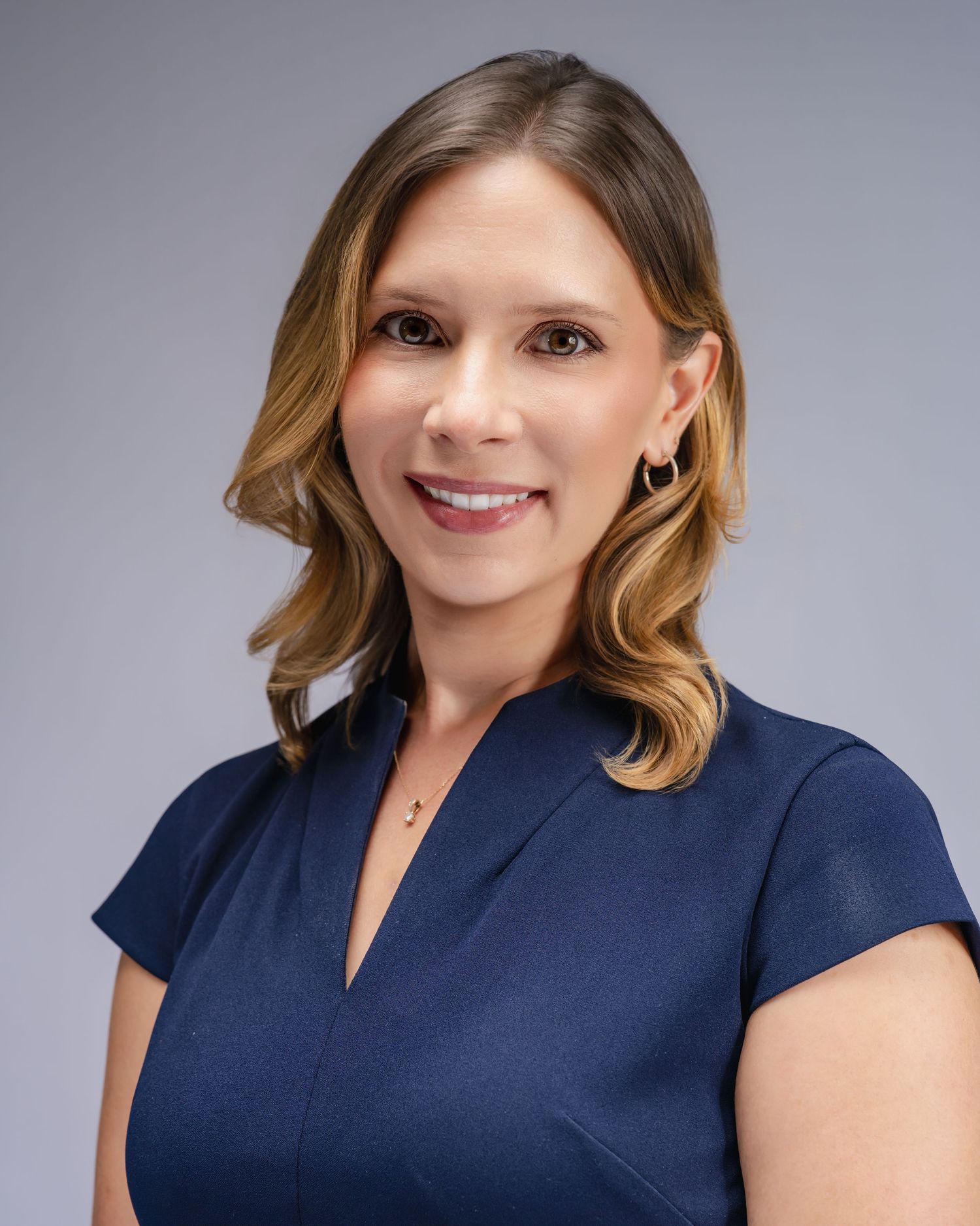 Professional headshot of a smiling woman with blonde highlights wearing a navy blue dress against a gray background.