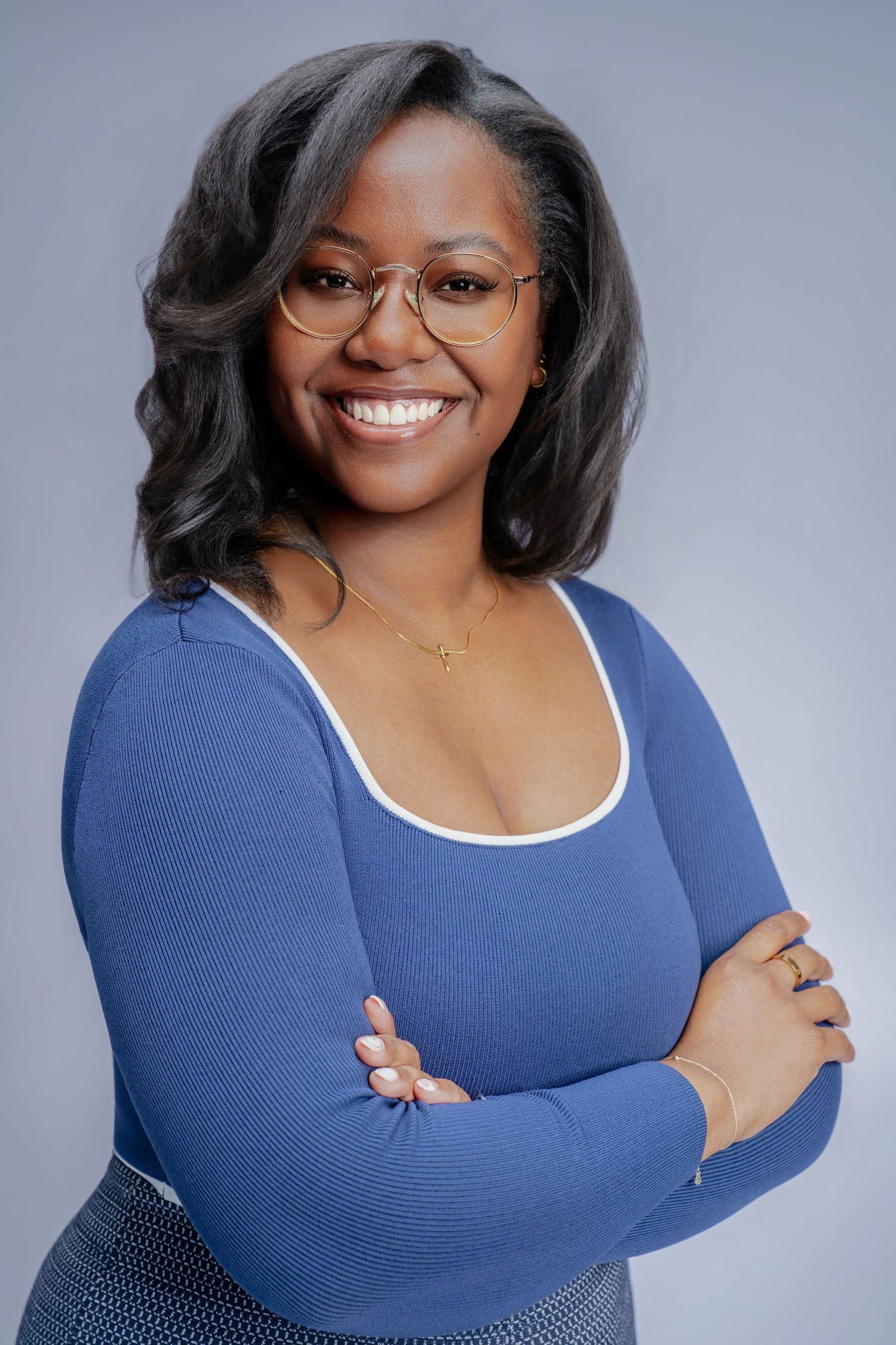 Smiling Black woman in blue top with glasses, arms crossed, studio portrait.