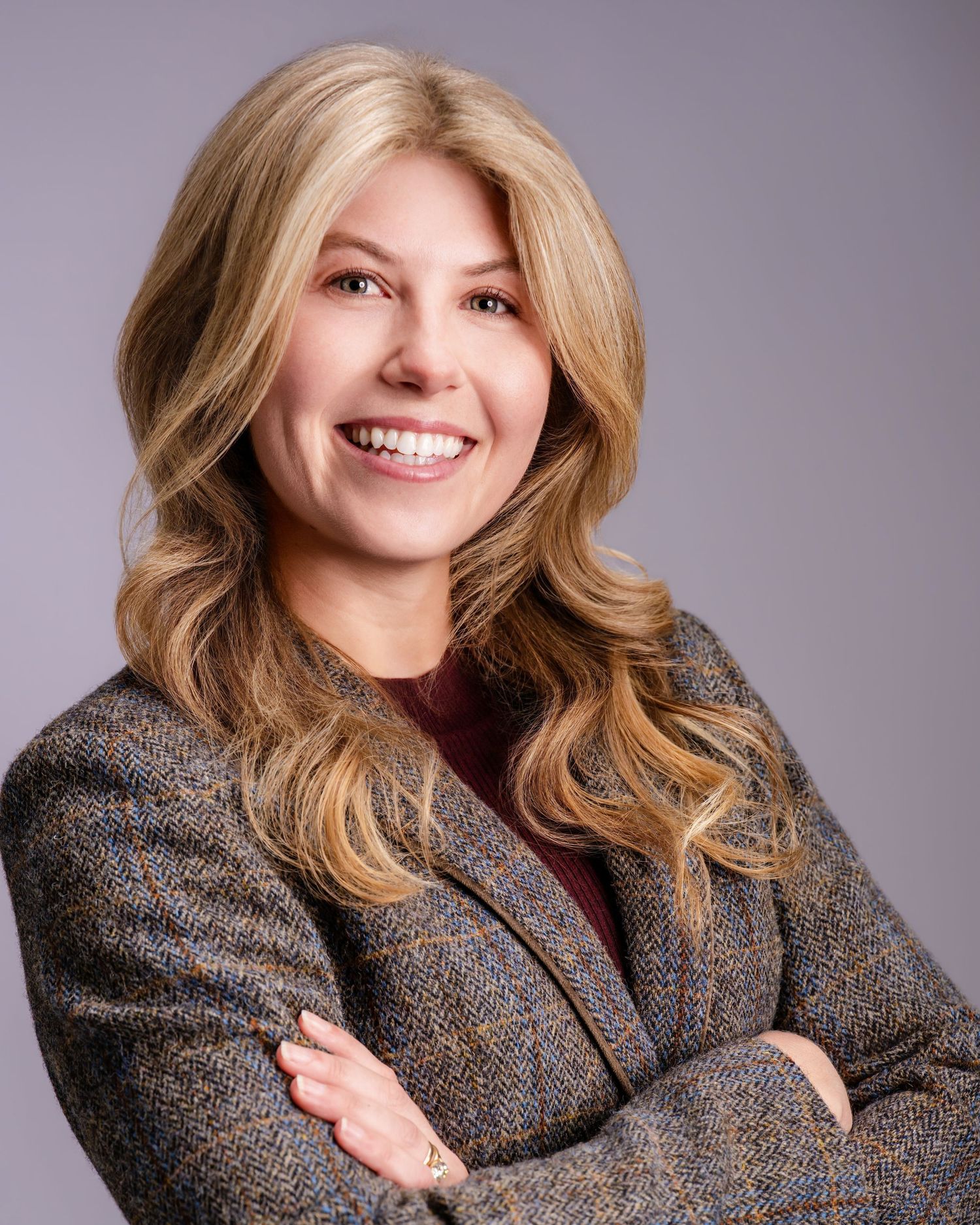 Professional headshot of smiling blonde woman in tweed blazer with arms crossed on gray background.