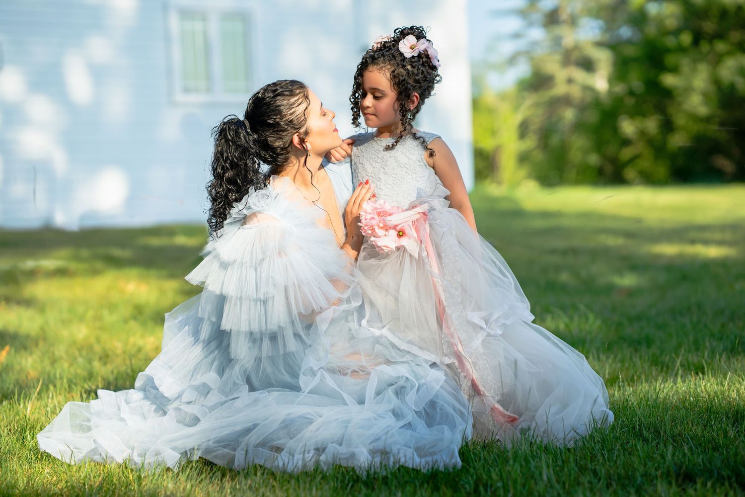 Woman and young girl in matching white tulle gowns sharing a tender moment on a green lawn.