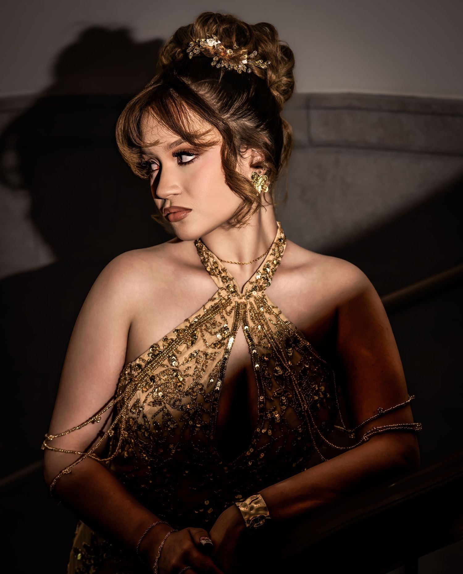 Young woman in gold embroidered gown with updo hairstyle and gold accessories in dramatic lighting.