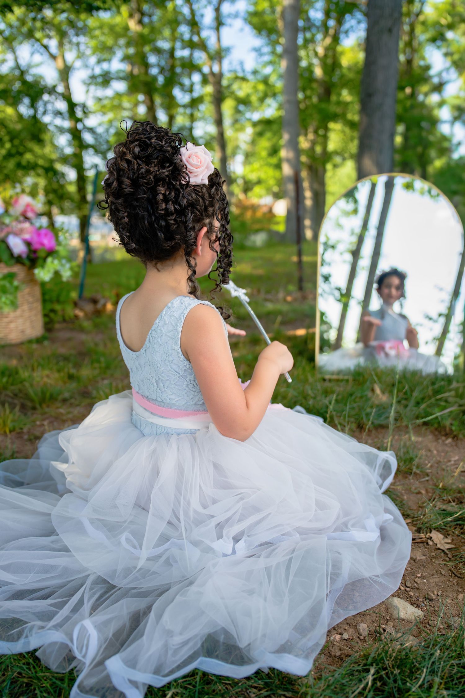 Young girl in a white tulle dress holding a wand, looking at her reflection in an outdoor mirror.