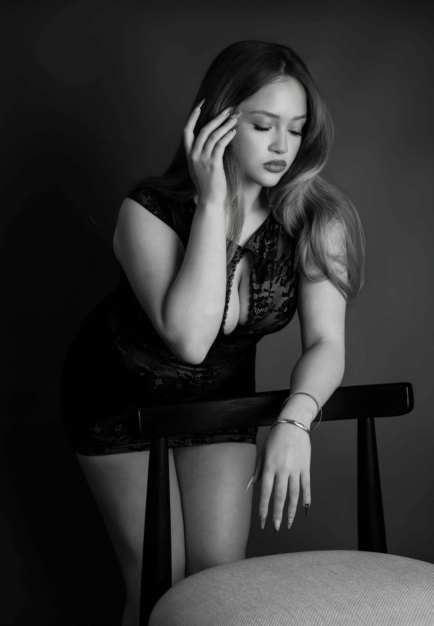 Black and white studio portrait of young woman in lace dress posing thoughtfully beside chair.