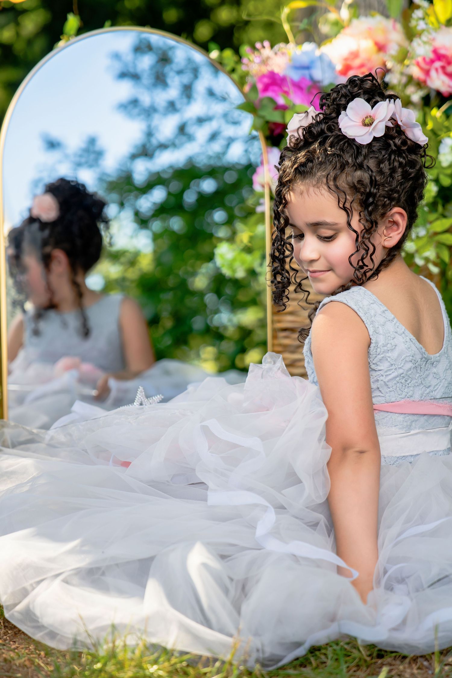 Young girl in a white tulle princess dress with flowers in her hair sitting beside a mirror outdoors.
