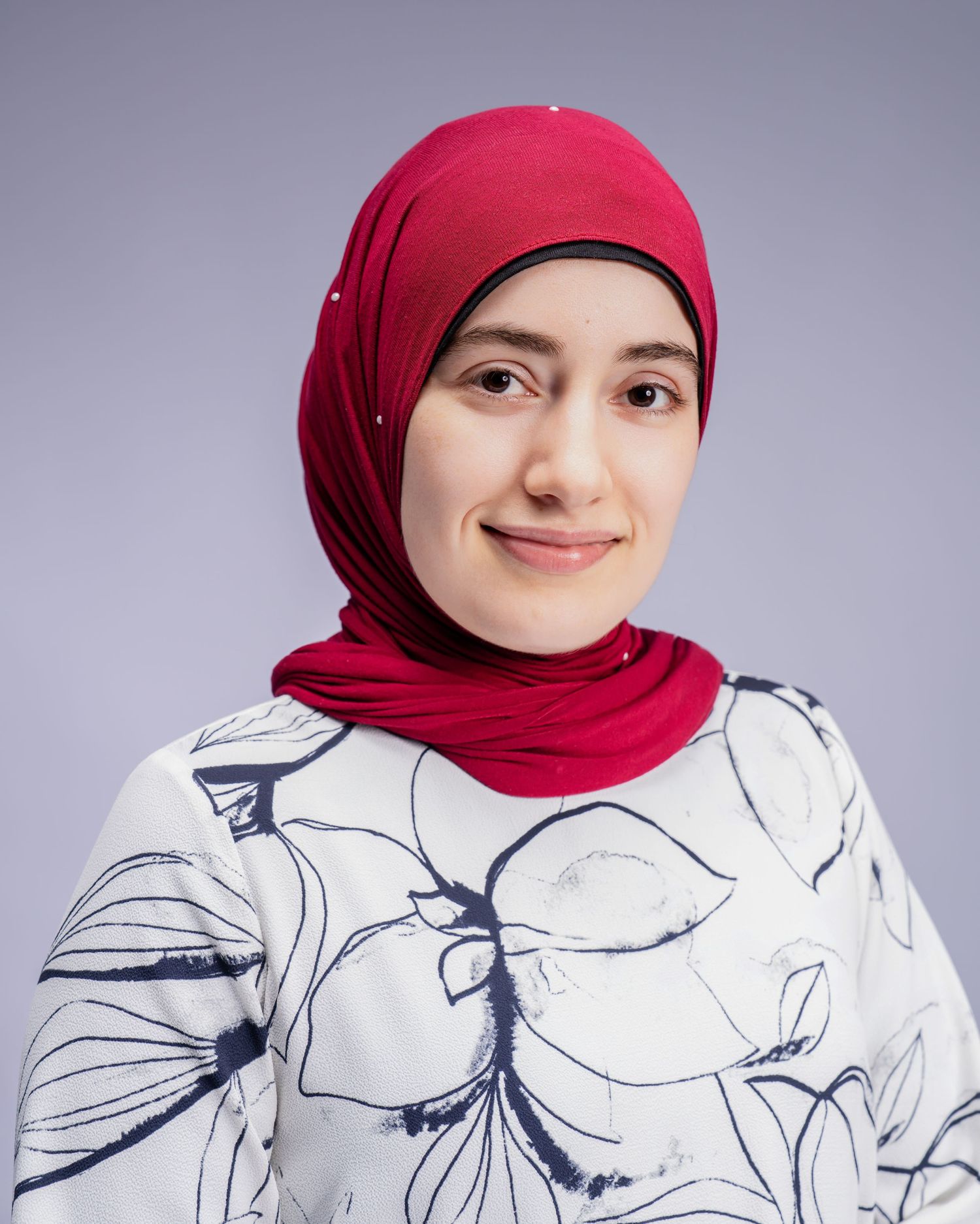 Smiling woman wearing a red hijab with floral white top, studio portrait.