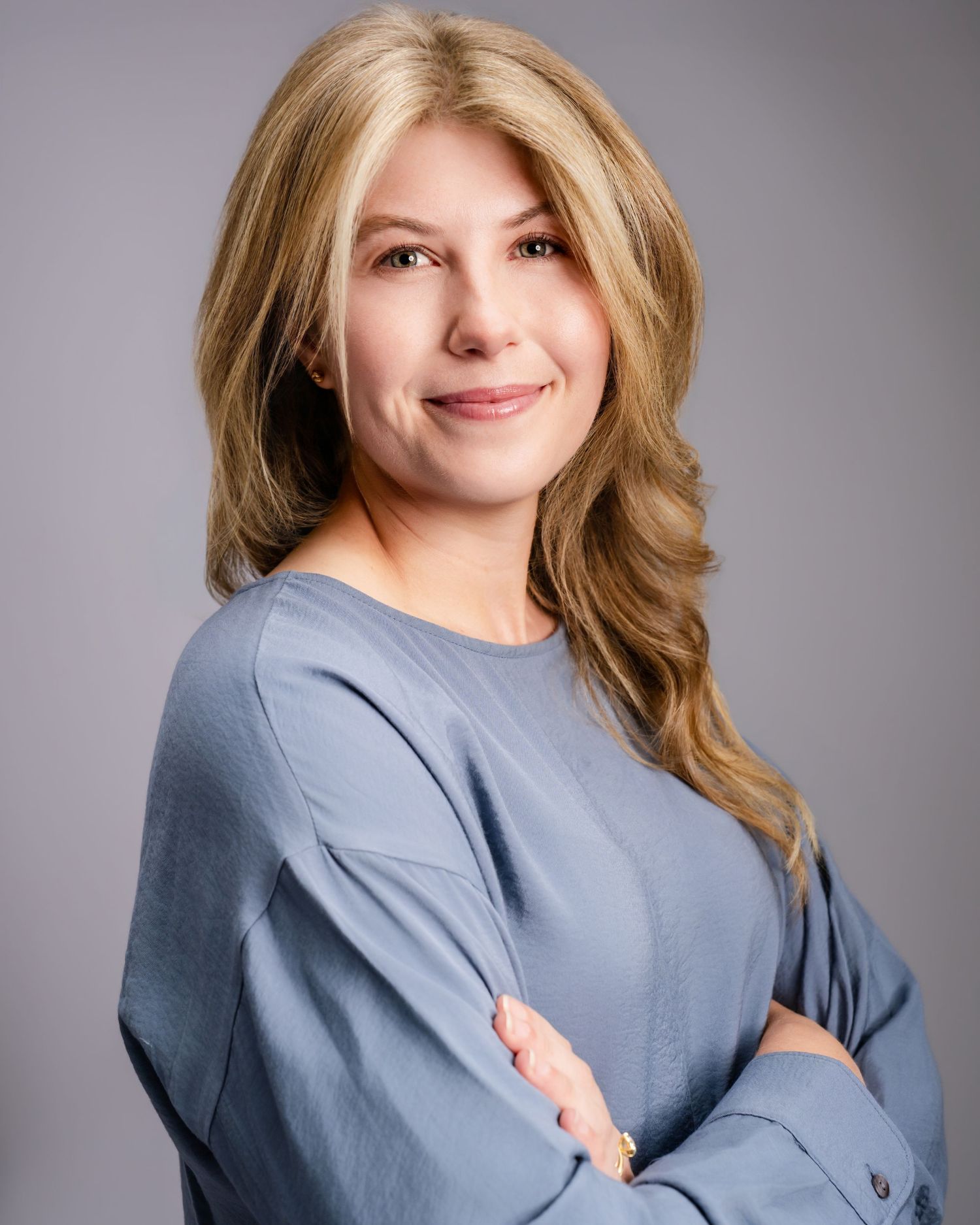 Professional headshot of smiling blonde woman in blue top with arms crossed on gray background.