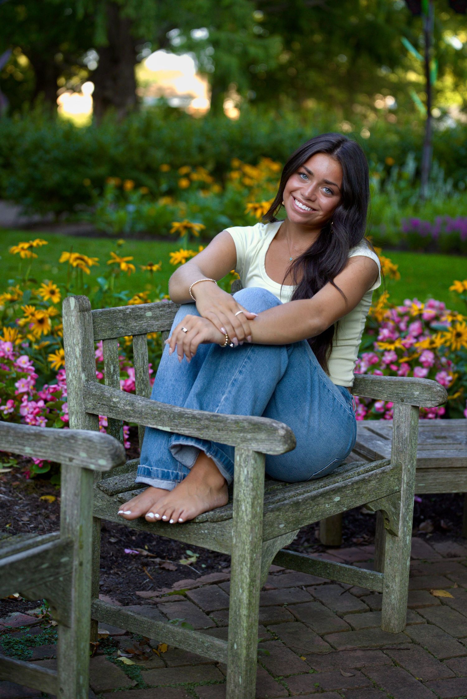 Smiling young woman sitting barefoot on a wooden park bench surrounded by colorful flowers and lush green trees.