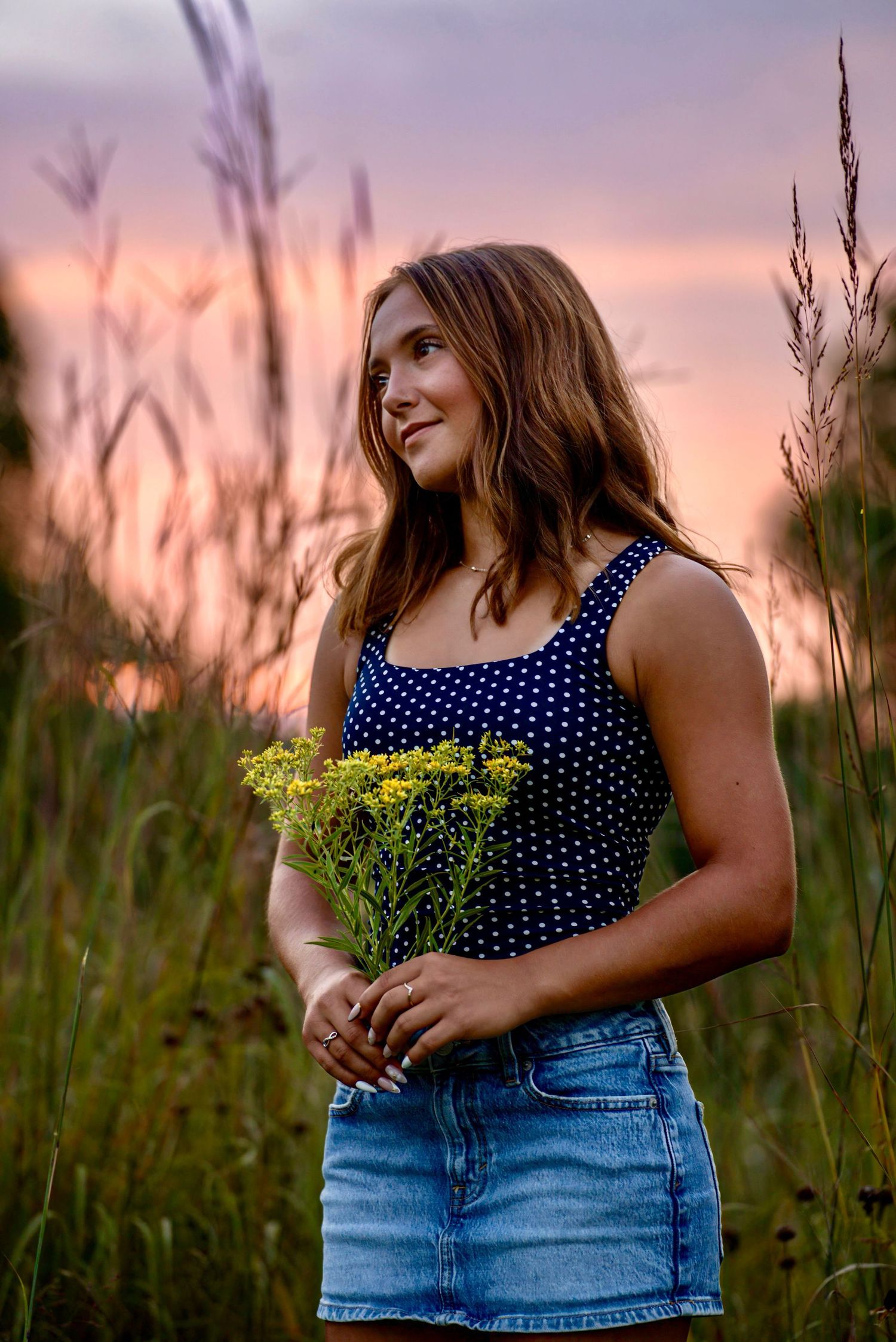 Young woman holding wildflowers in a field at sunset, wearing a navy polka dot top and denim skirt.