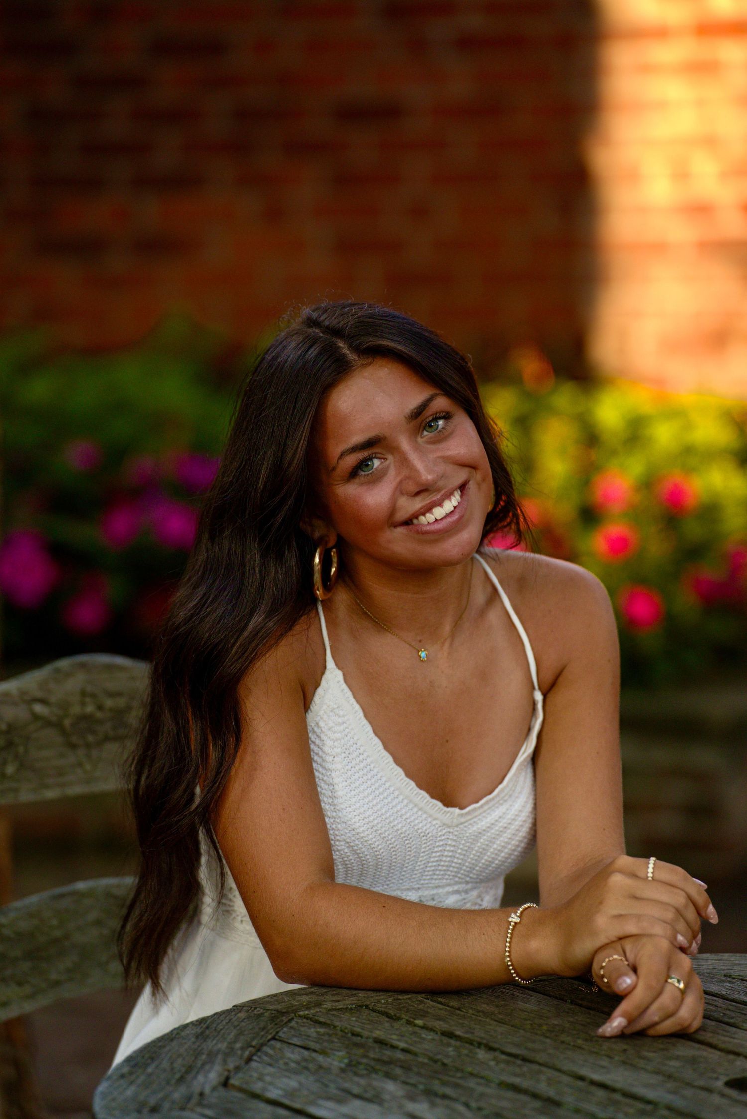 Smiling young woman with long dark hair wearing a white dress, seated at an outdoor table surrounded by colorful flowers.