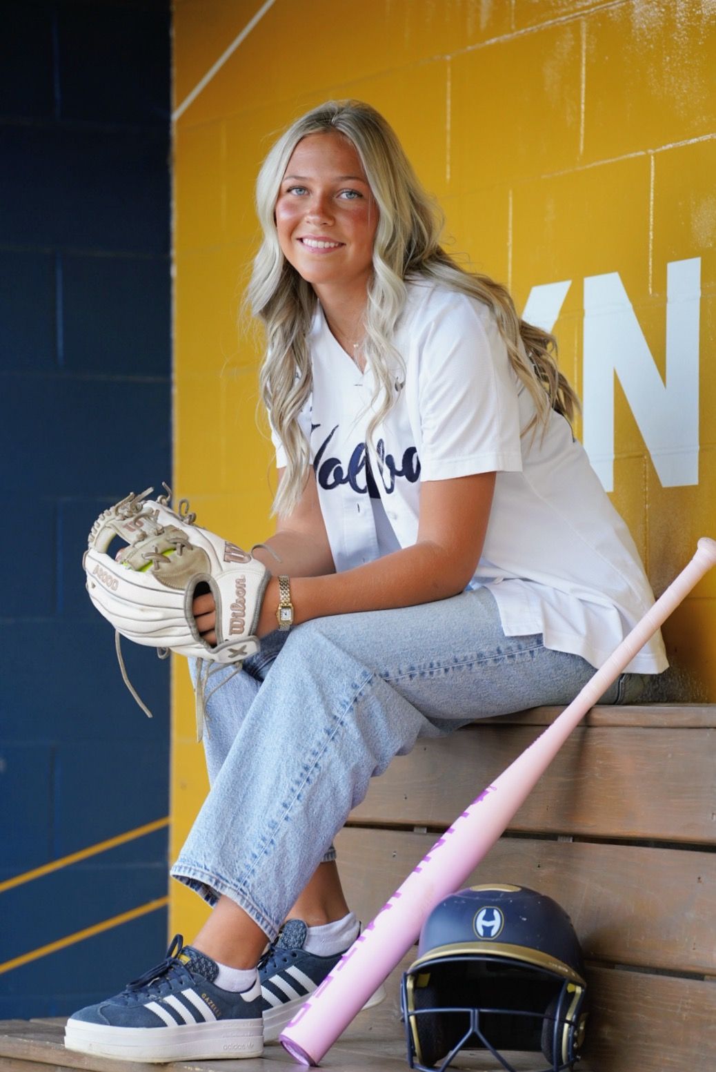 Young blonde softball player smiling in dugout, holding glove, with pink bat and helmet nearby.